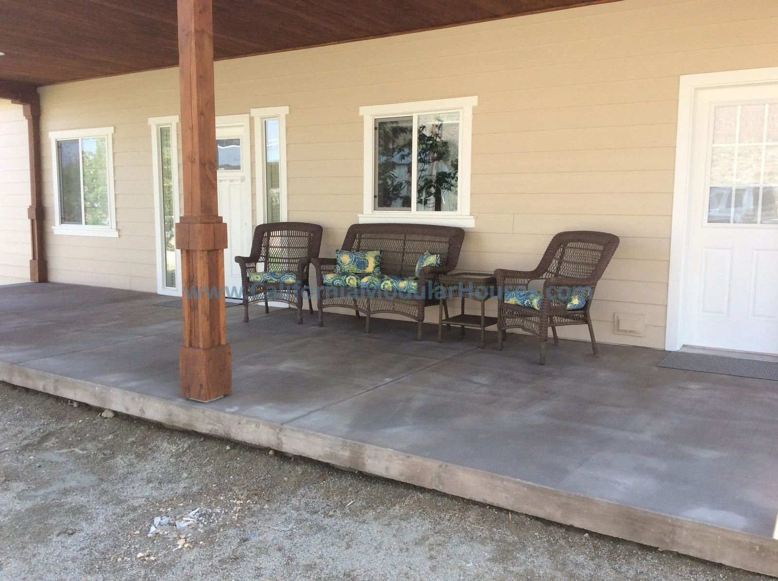 A porch with a concrete floor, beige siding, and white trim windows and door. There are three dark brown woven chairs with blue and yellow cushions, and a tall wooden support post.