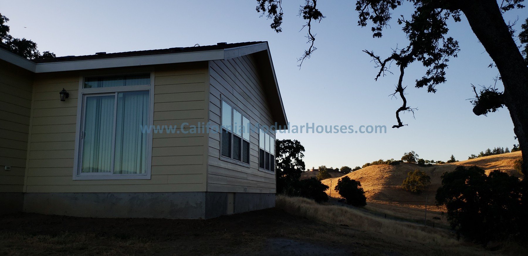 A house with beige siding and sliding glass doors on a sloped yard, with a landscape of rolling hills and trees in the background during sunset.  California Modular, Prefab Modular Homes California.