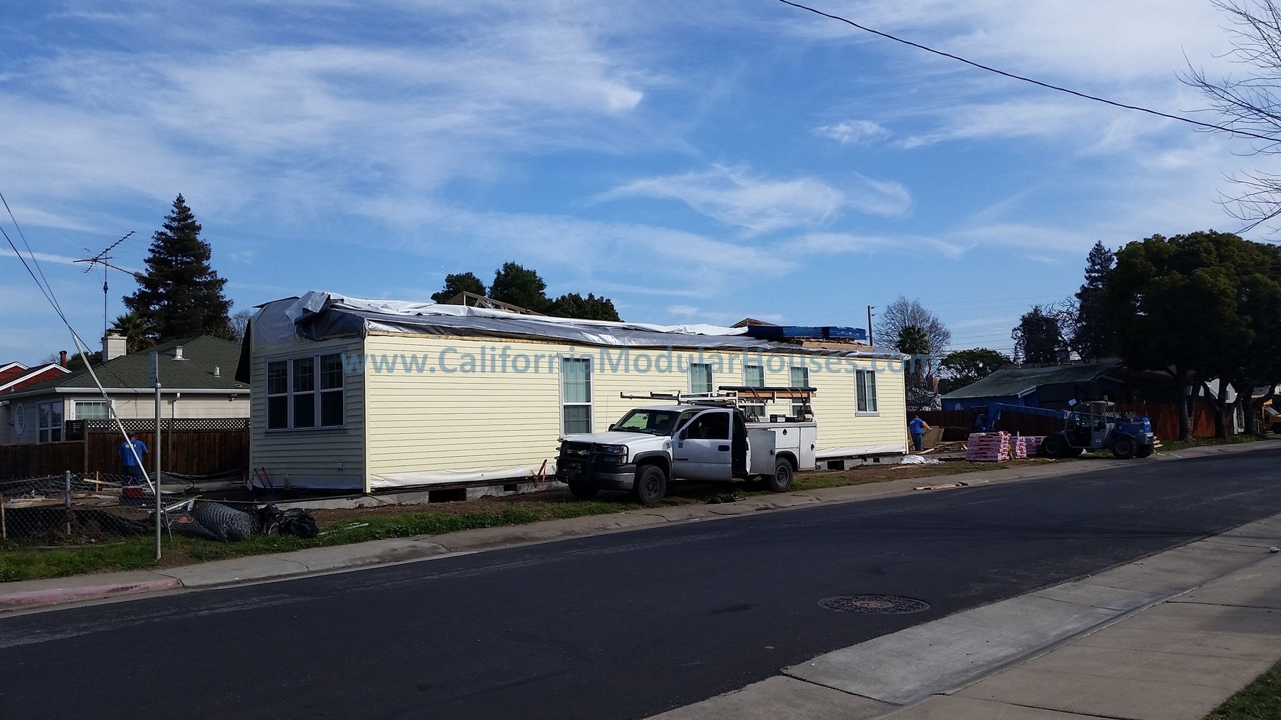 Under construction of a single-story house, with a white utility truck parked outside, and construction materials like pink insulation stacked nearby. The house has beige siding and several windows, with some construction materials visible on the roo