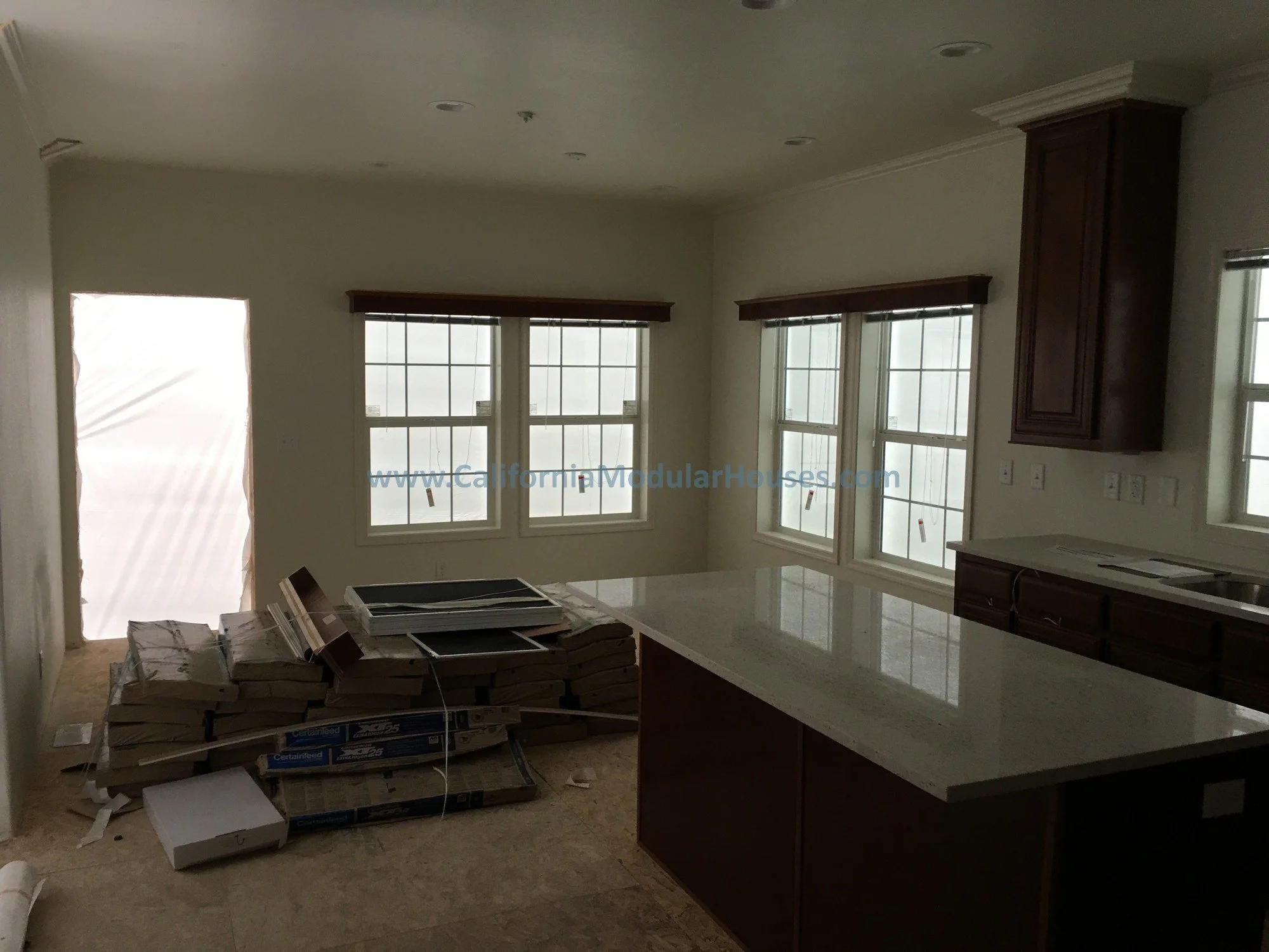 Empty kitchen with beige walls and countertops, dark wood cabinets, and large windows covered with protective film, with packages of flooring and tools on the floor.