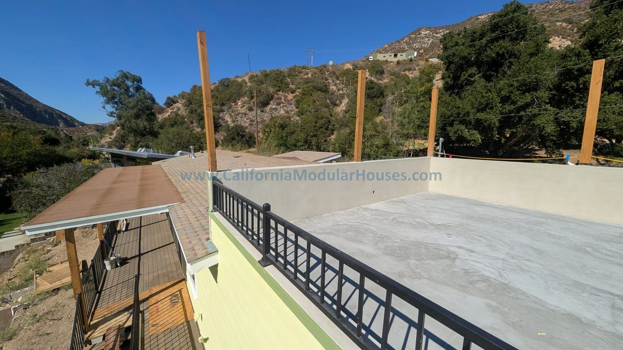 View of a rooftop patio under construction with a black metal railing, wooden posts, and a concrete floor, overlooking hills with trees and houses in the background.