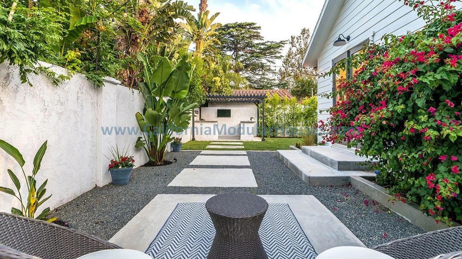 A backyard patio with a white house on the right, a white wall on the left, and a lawn in the background. There are plants and flowers along the sides, a black outdoor stove or grill, and a wicker table with chairs in the foreground.