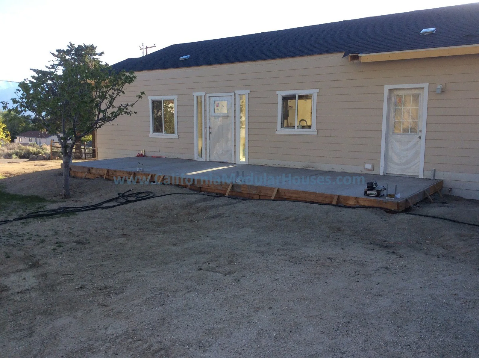 Construction site of a beige house with a newly poured concrete patio and a single tree on the left side.