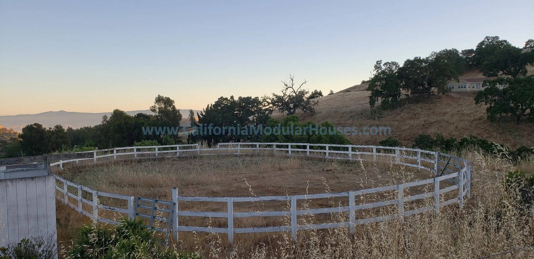 A circular horse riding arena with white fencing surrounded by dry grass, trees, and hills under a clear sky at sunset.   Bay Area Prefab Homes, California Modular Home, 