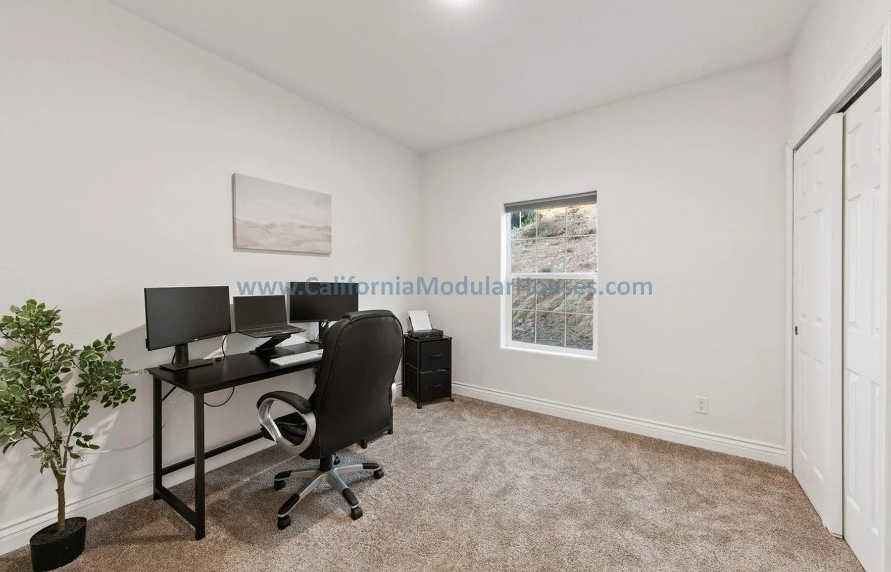 A simple home office with a desk holding three computer monitors, a laptop, and a keyboard, a black office chair, a small black filing cabinet, a potted plant, a window showing outdoor terrain, white walls, and a closed closet door.