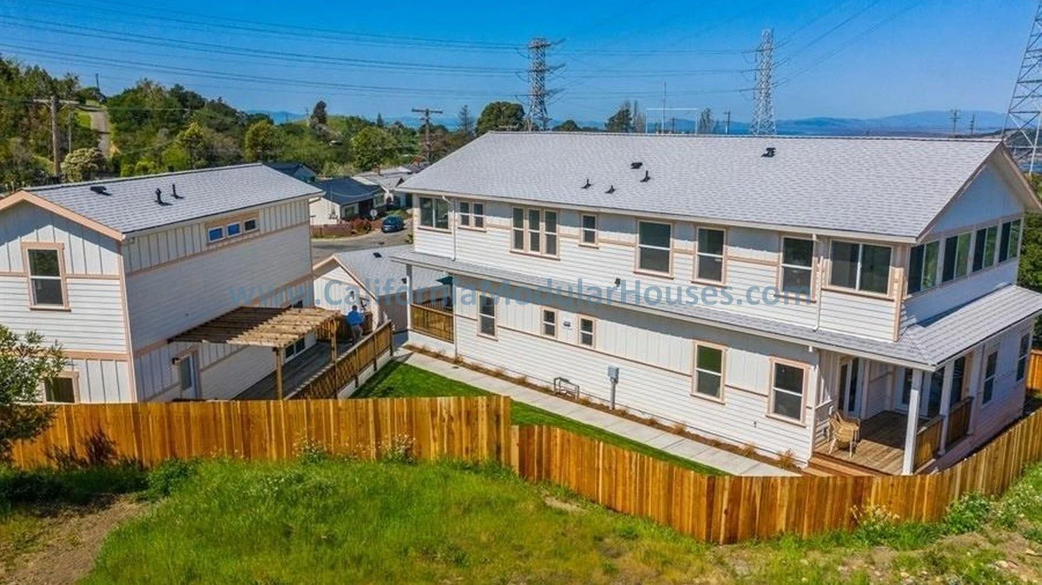 An aerial view of two white suburban houses with gray roofs, a fenced backyard, and power lines in the background.  Single Family Residence.  Two Story and an Accessory Dwelling Unit.  Prefab Modular.  