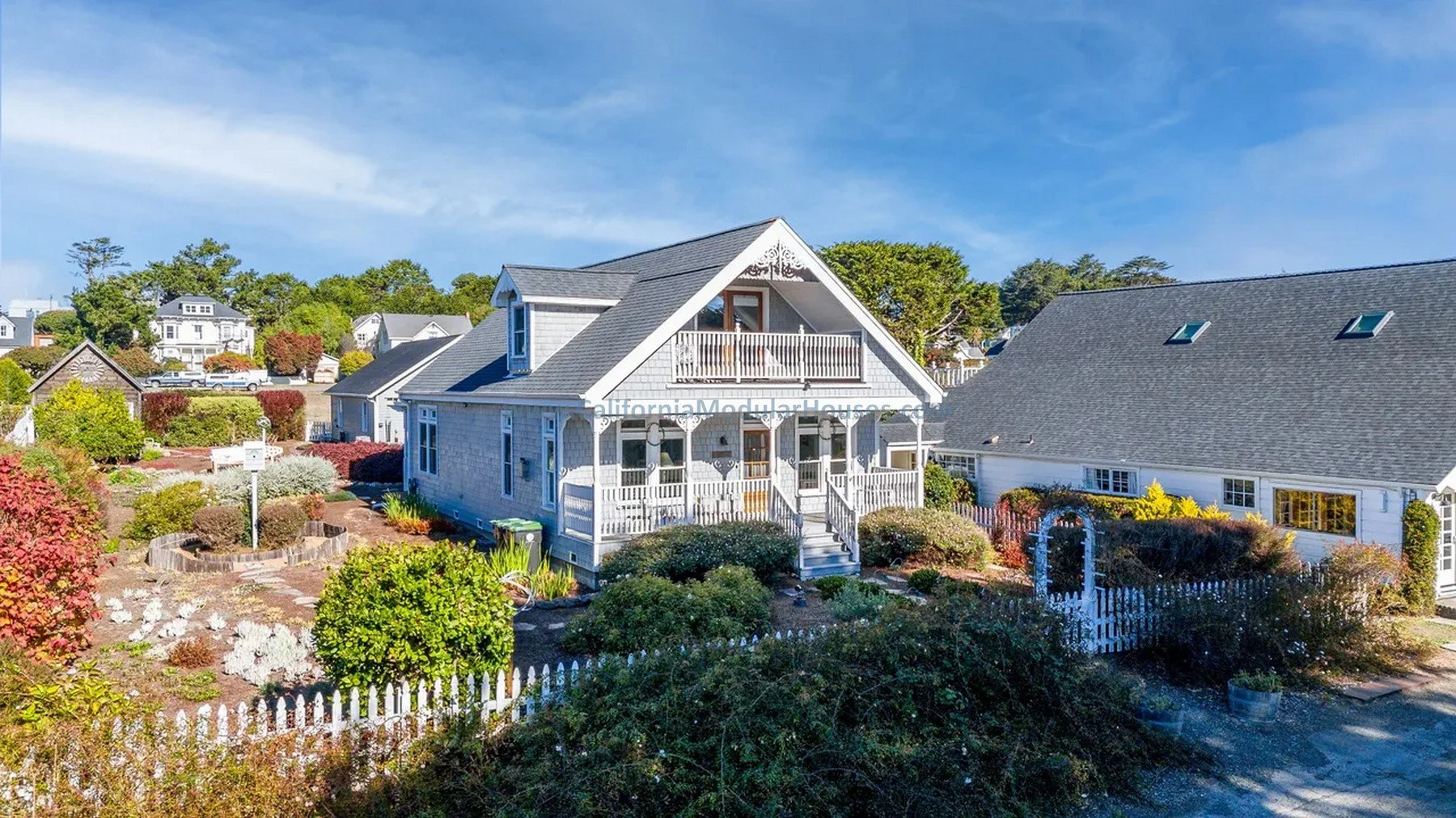 A white two-story house with a front porch, balcony, and gray shingle roof surrounded by a colorful garden with shrubs and flowers.  Modular Homes in California, Pre-Fabricated Homes.