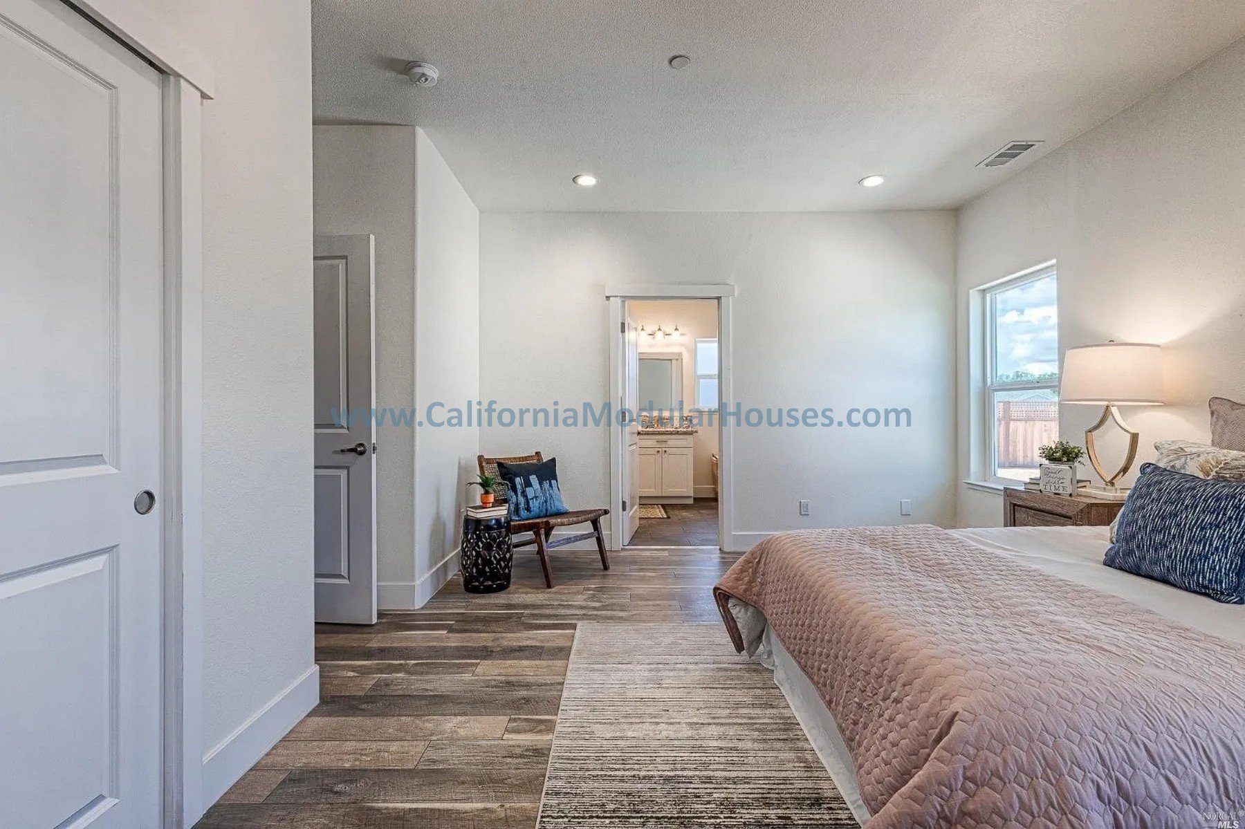 A bedroom with a bed on the right, a nightstand with a lamp and plant, two windows, a chair with a pillow, and an a door leading to a bathroom.