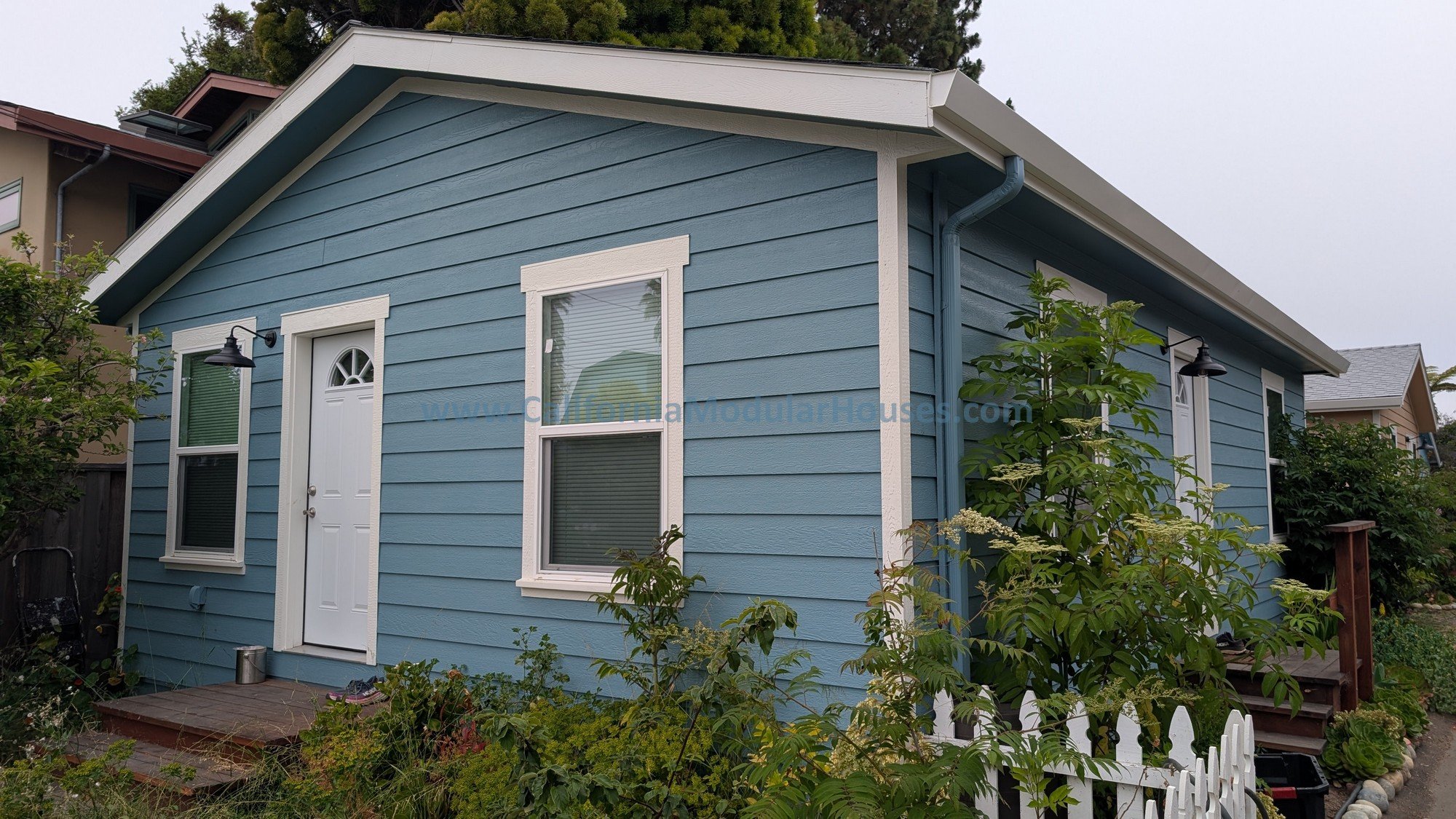Blue house with white trim, two windows, front door, outdoor lights, surrounded by garden, shrubs, and plants.
