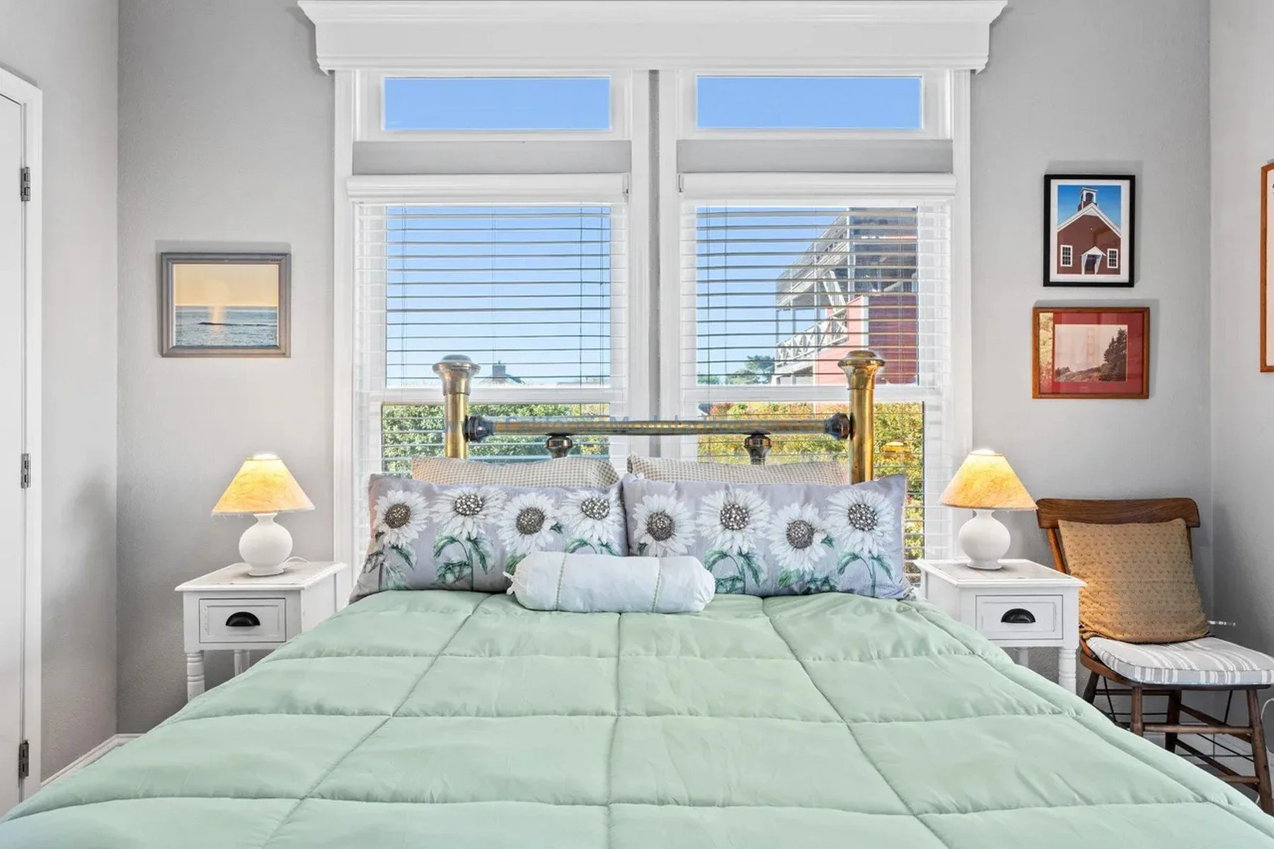 A bedroom with a bed, green comforter, sunflower-patterned pillows, two white nightstands with lamps, and windows with blinds showing a blue sky and nearby building.  Mendocino Historic area.  Modular.  Pre-fab.