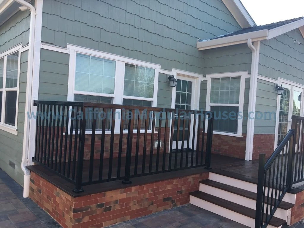 Exterior view of a green house with a small wooden deck, black railing, and brick base, with steps leading up to a door.