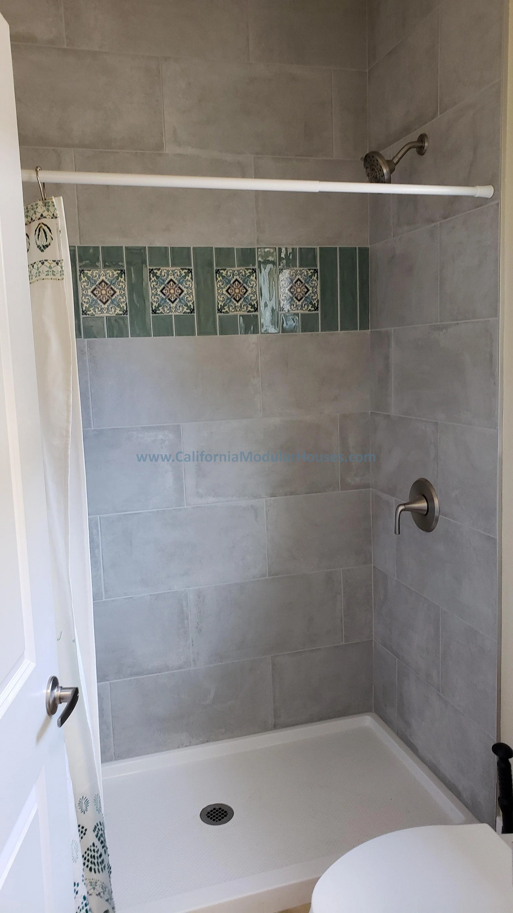 A walk-in shower with gray tiles, green decorative tiles, a showerhead, a curved shower rod, and a shower drain. A partial view of a white door and a curtain is visible.  Interior Modular Prefab ADU Photograph