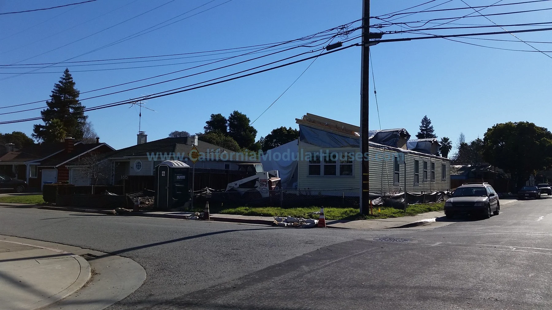 Construction site with a house under renovation, construction materials, a portable toilet, and parked cars on a street under a clear blue sky. Bay Area Prefab Homes, Modular Homes California,