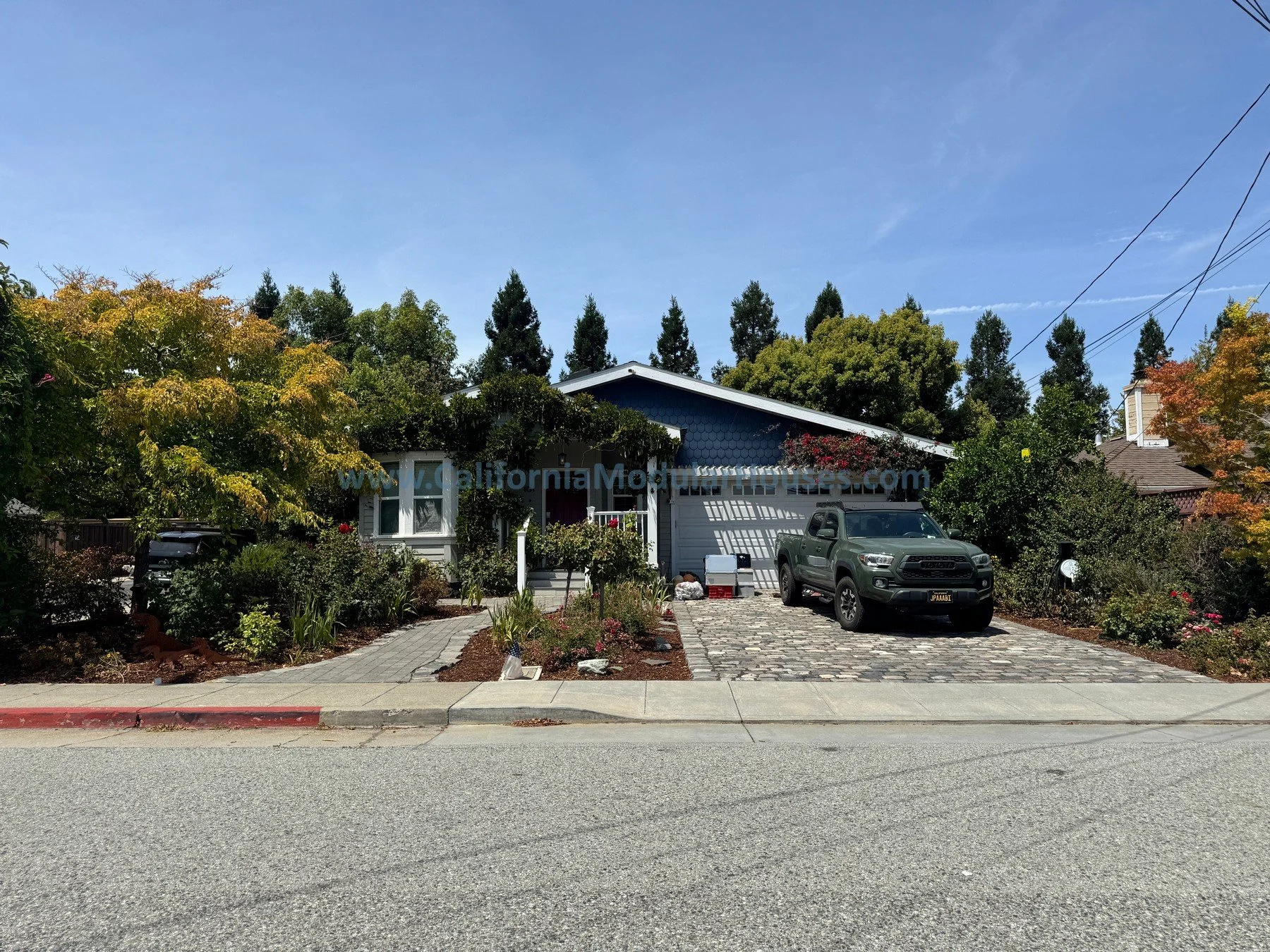 A suburban house with a blue exterior and a sloped roof, surrounded by green trees and bushes. A gray pickup truck is parked in the driveway on a cobblestone path. The street, curb, and sidewalk are visible in the foreground, with a red-painted curb.