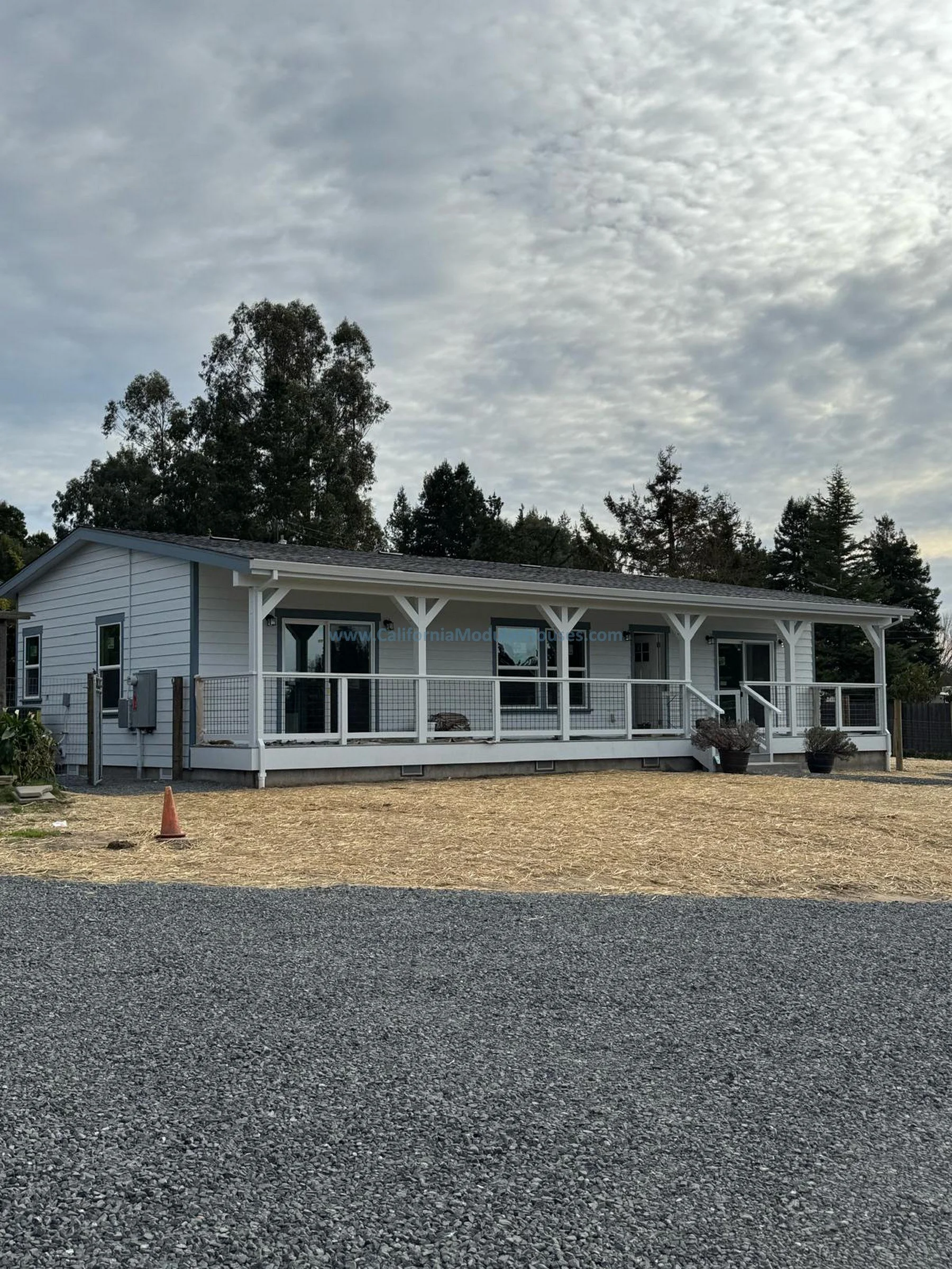 A single-story white house with a front porch, surrounded by trees and a mostly gravel yard, under a cloudy sky.  Accessory Dwelling Unit Prefab Modular Home for Sebastopol, CA.  Modular ADU California.  Sonoma County.