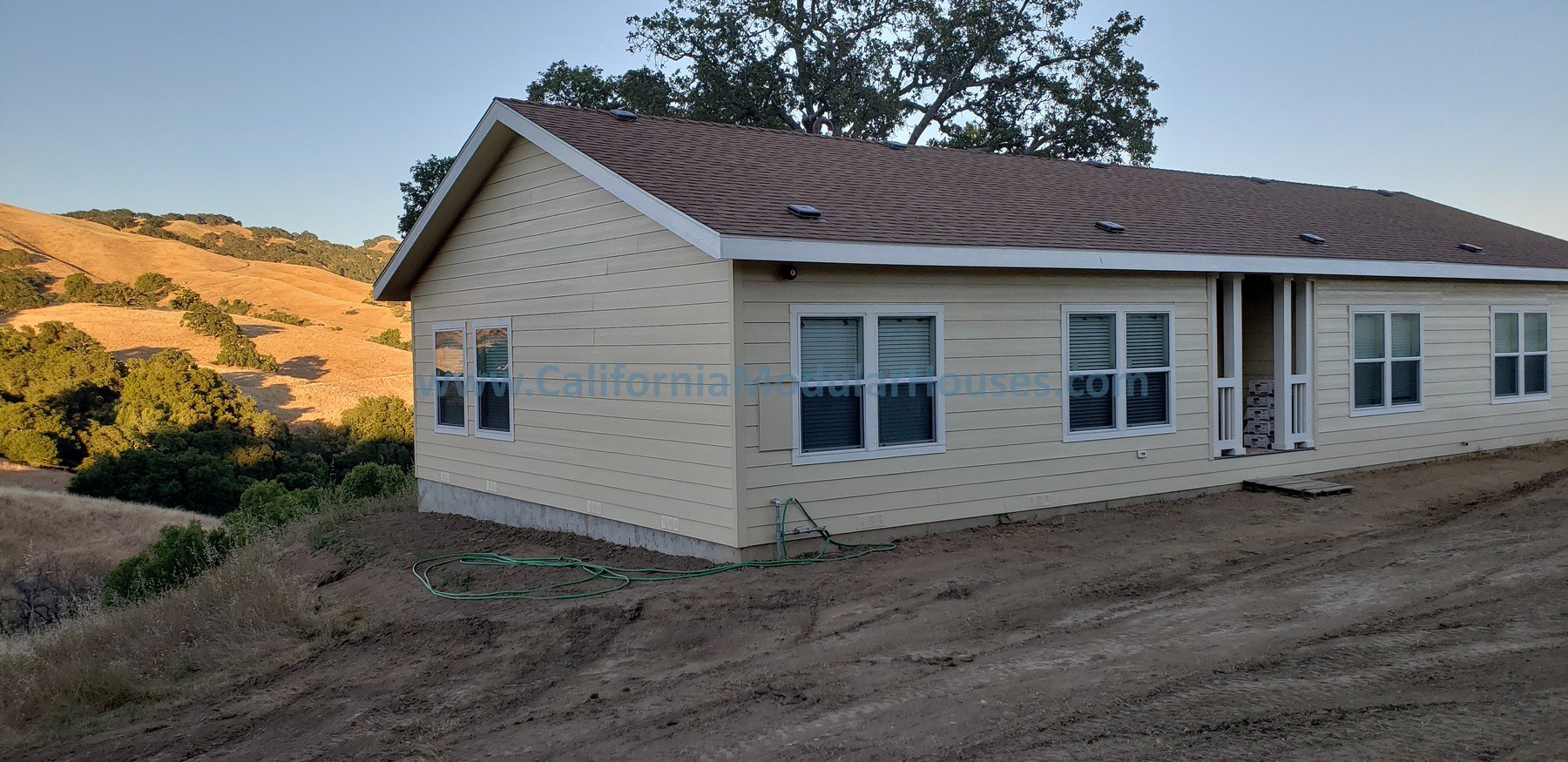 A newly built house on a dirt lot, with a light yellow exterior and a brown roof, surrounded by rolling hills and trees in the background. California Modular Houses,  Bay Area Prefab Modular Home, Modular Homes,