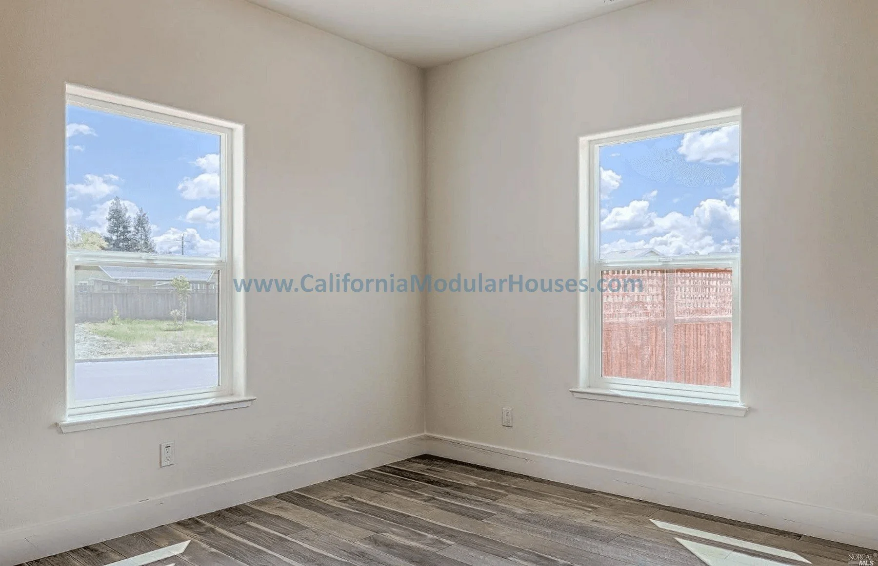 Empty room with white walls, two windows showing a blue sky with clouds, wood flooring, and baseboards.