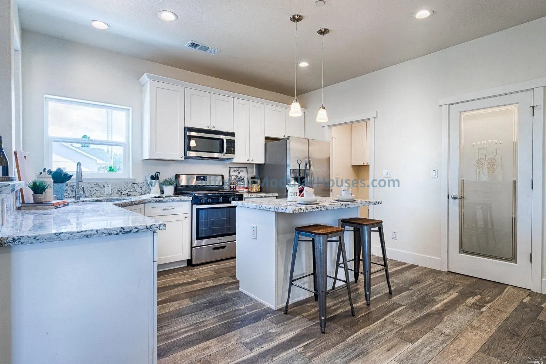 Modern kitchen with white cabinets, stainless steel appliances, marble countertops, and wood flooring. Includes a small breakfast island with two stools and decorative items.