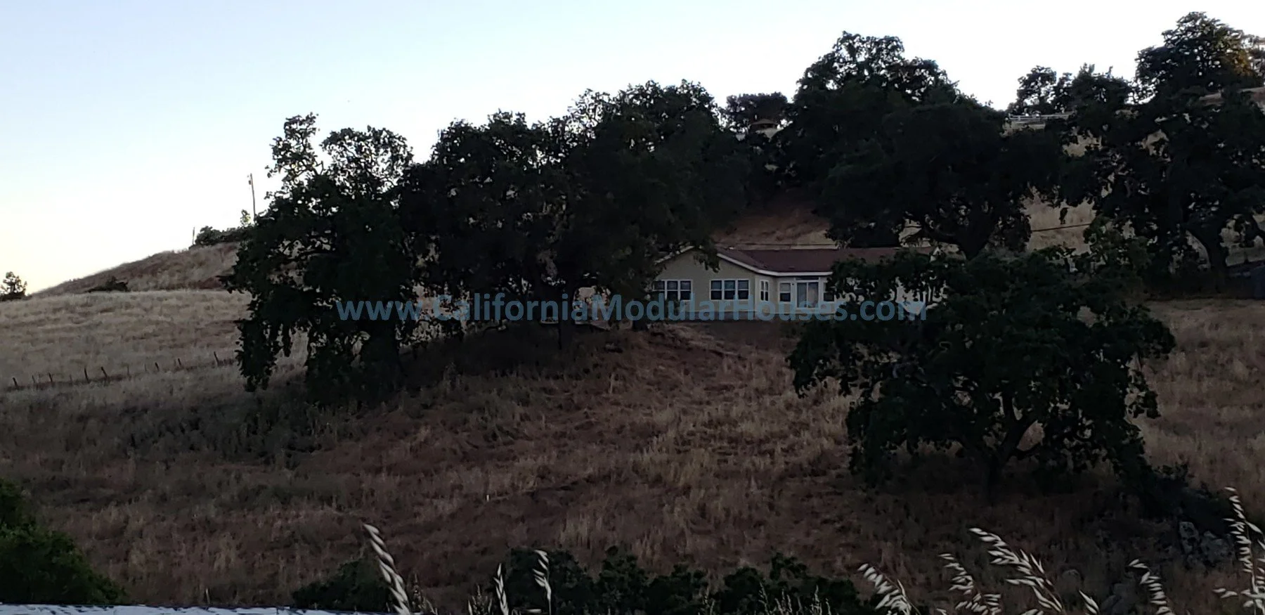 House on a hillside partially obscured by large trees, with dry grass and a fence in the foreground, and a clear sky in the background. Modular Homes California, Modular Homes in California, San Jose, CA.  