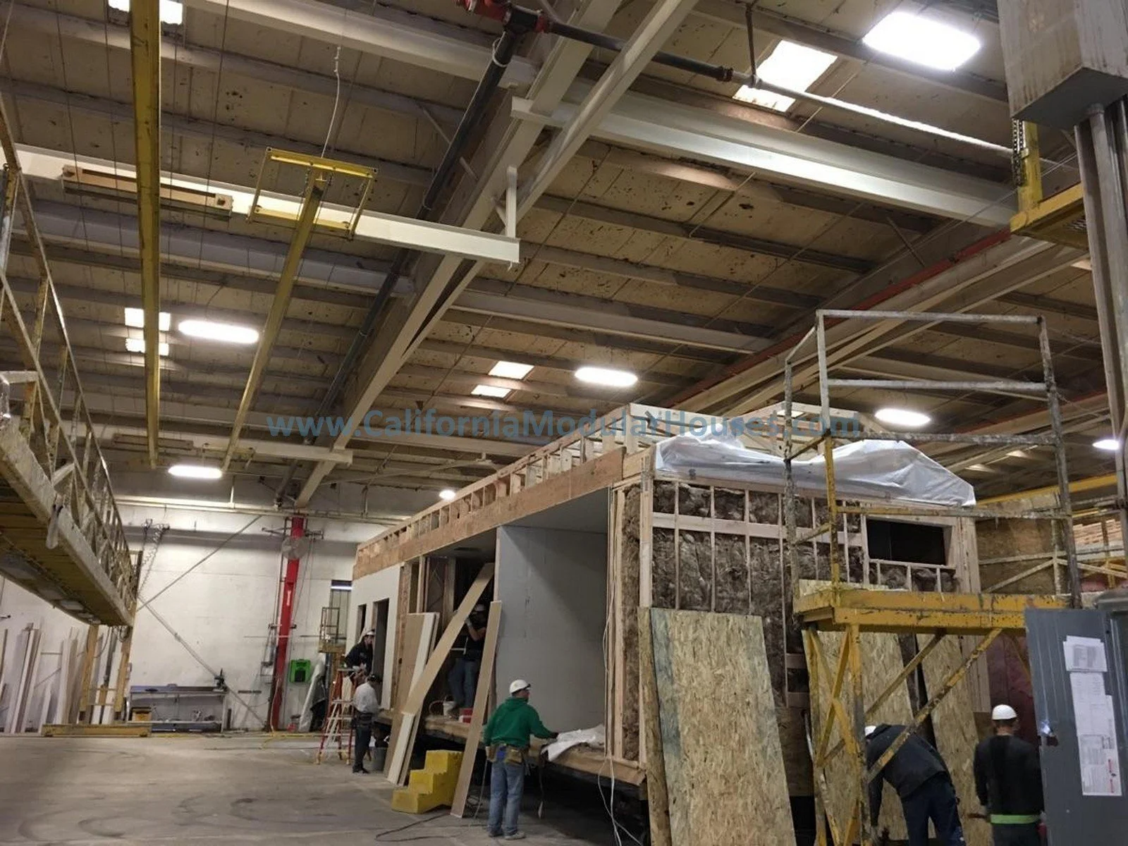 Workers building a small structure inside a large factory with scaffolding and construction materials.  Factory Built Housing.  