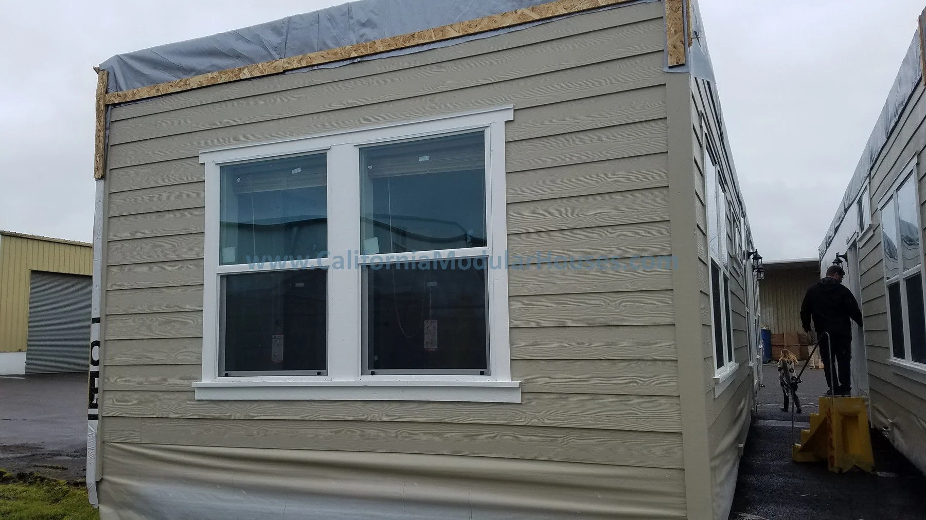 View of a newly constructed beige modular home with multiple windows and white trim on a cloudy day, with workers visible on the right side.  California Modular Home,  Modular Homes, Modular Homes California, Modular Homes in California,