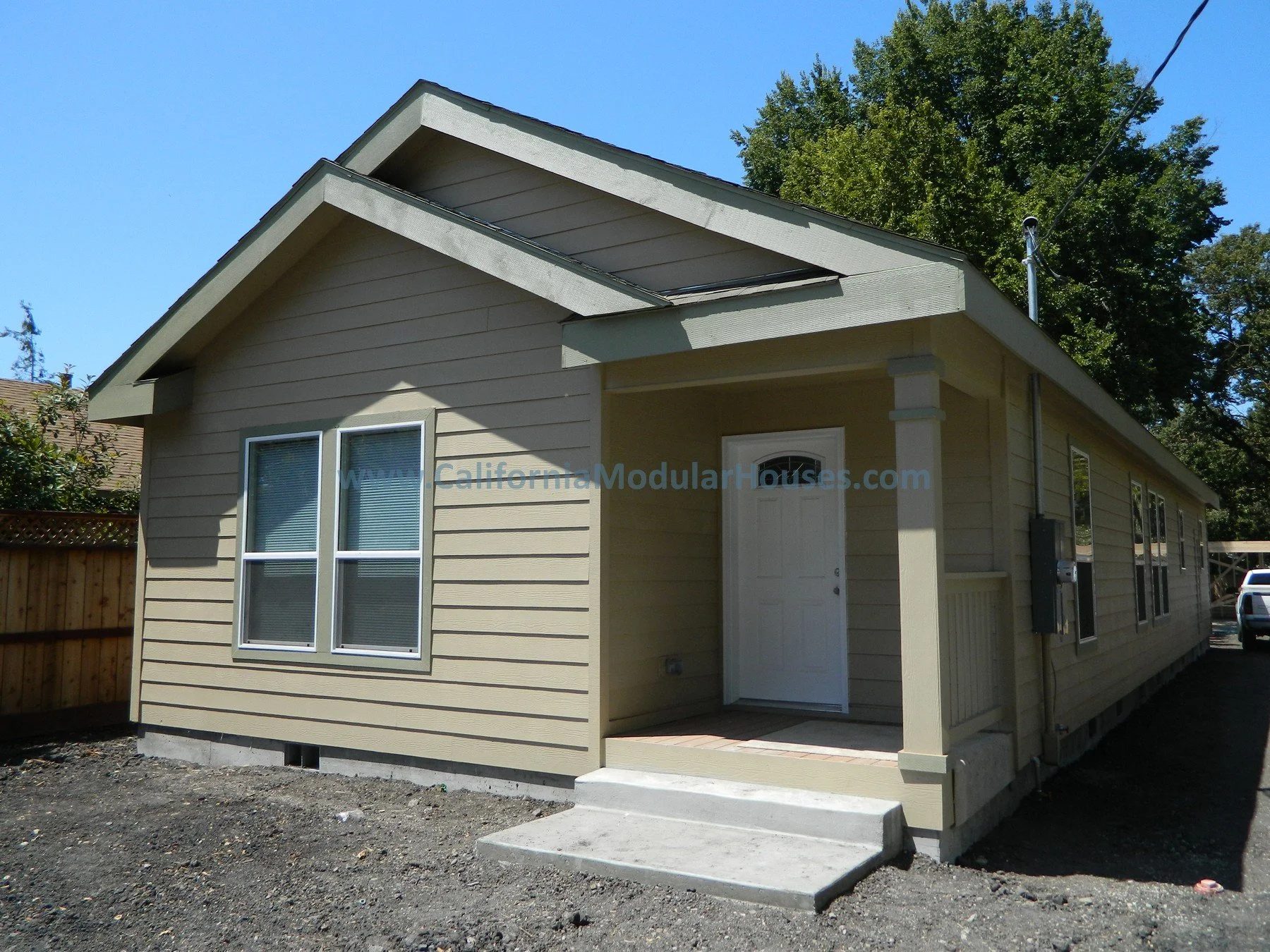 A newly built beige modular house with a small front porch, white door, and multiple windows, on a gravel lot with a wooden fence and trees in the background.