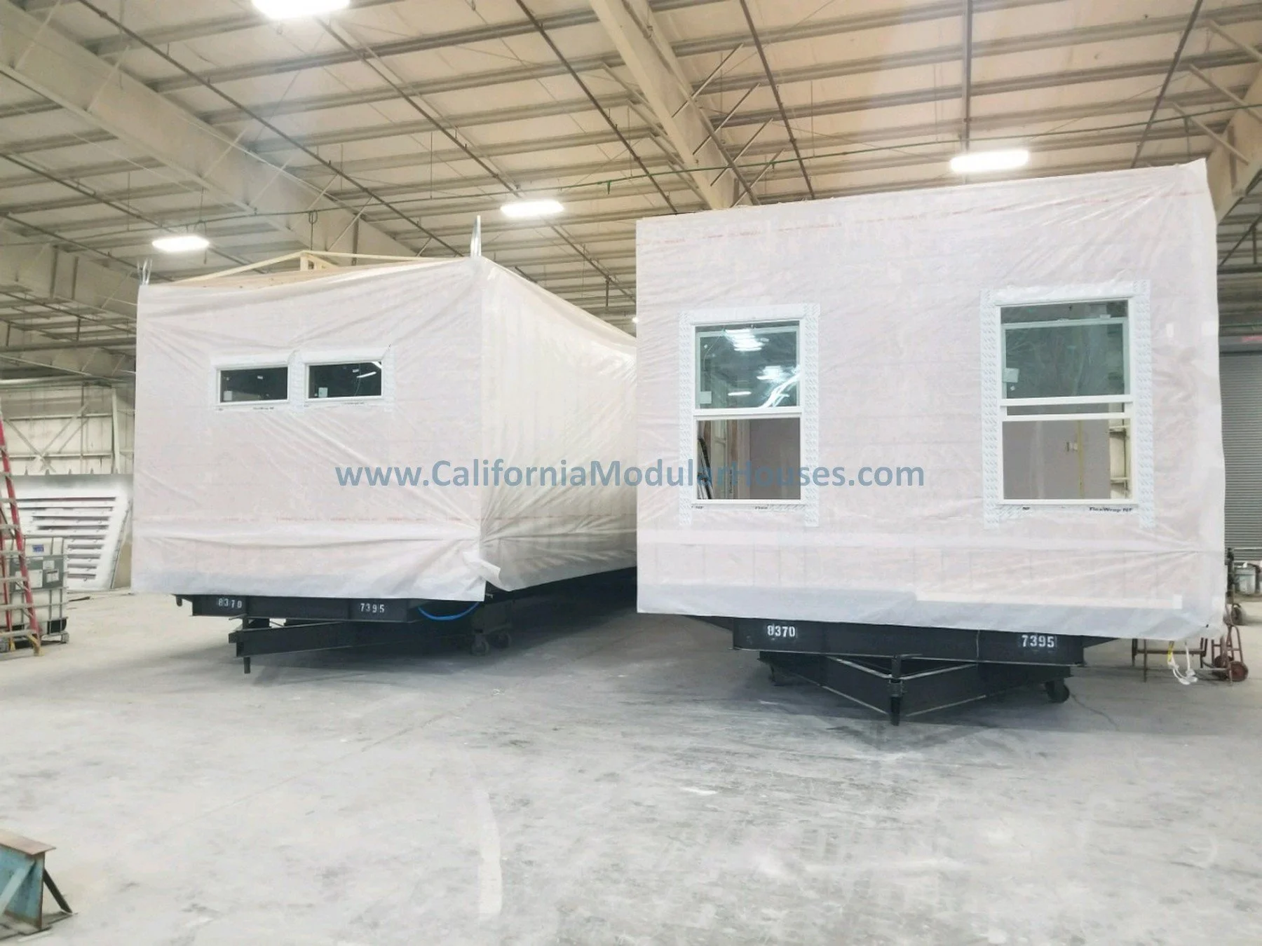 Two prefab modular buildings covered in white protective wrapping inside the factory, with windows visible on the structures.  Two-Story Modular Home.  City of Santa Barbara, Santa Barbara County, CA.  Modular Homes.