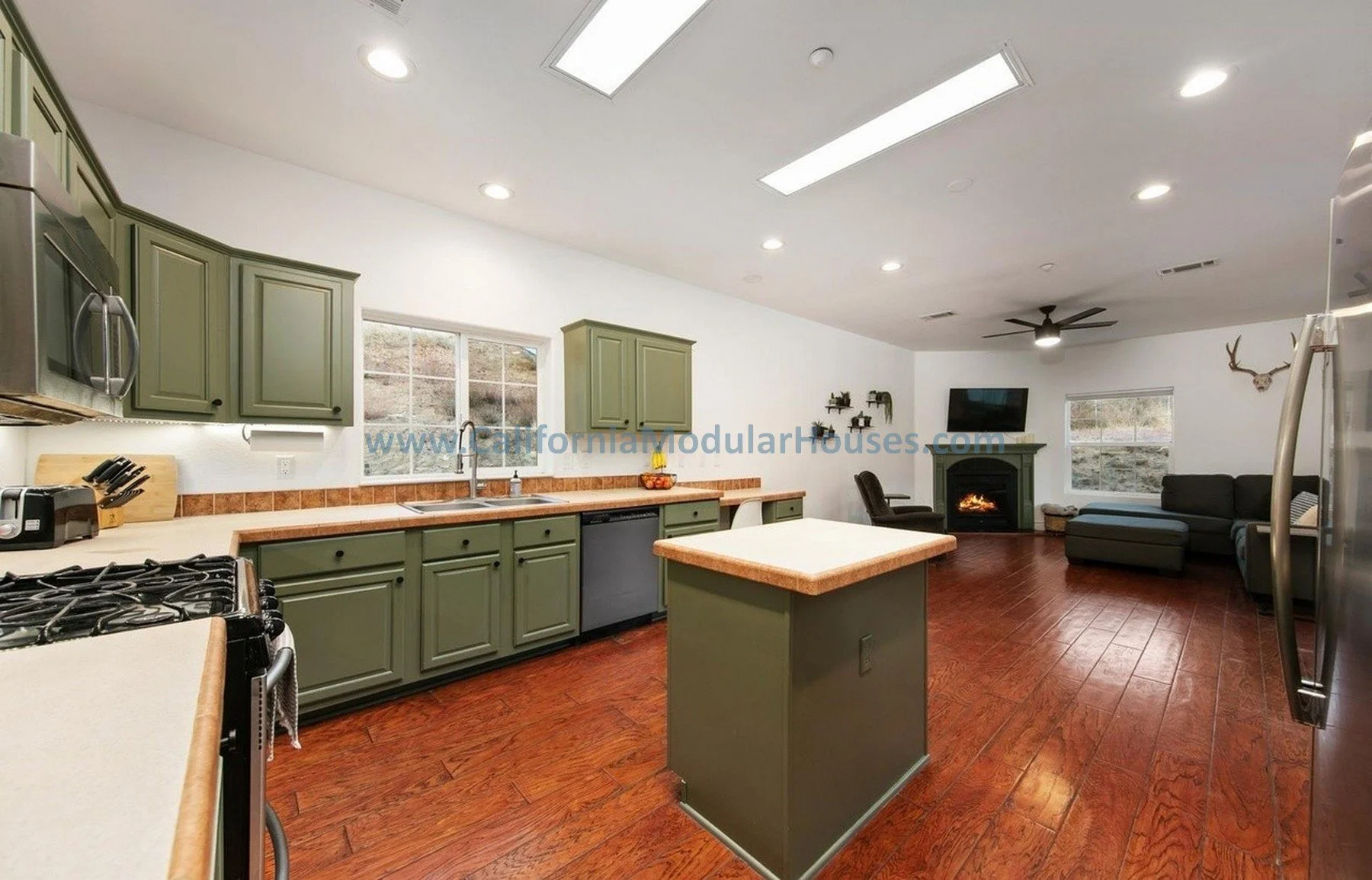 Open concept kitchen and living room with green cabinets, a center island, wooden floors, a fireplace, a TV, a black sectional sofa, and windows showing a hillside outside.