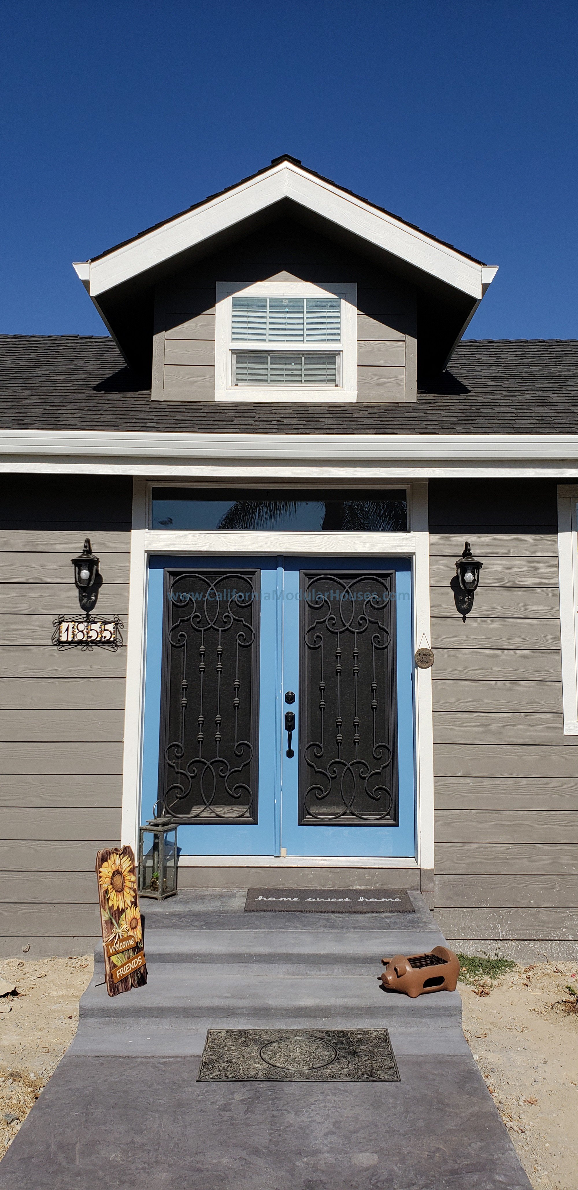 Front door of a modern house with blue painted frame and black wrought iron security doors, surrounded by gray siding, decorative concrete steps and a dormer above for the best appearance and a blue door painted to "pop".  