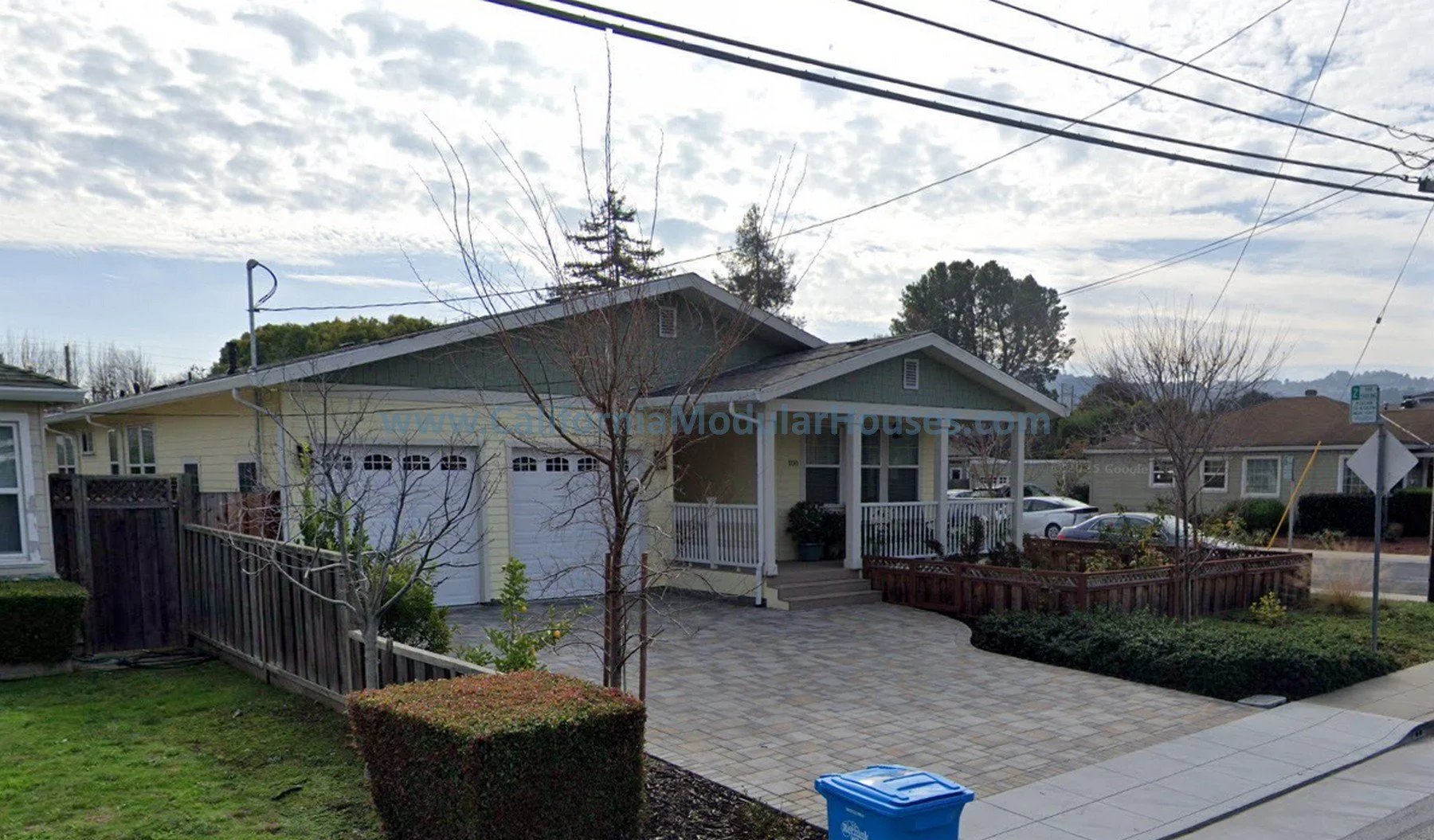 A suburban house with a yellow exterior, white garage door, and front porch with potted plants. The yard features a small tree, bushes, and a lawn with a small hedge. There are parked cars nearby, telephone wires overhead, and a partly cloudy sky.