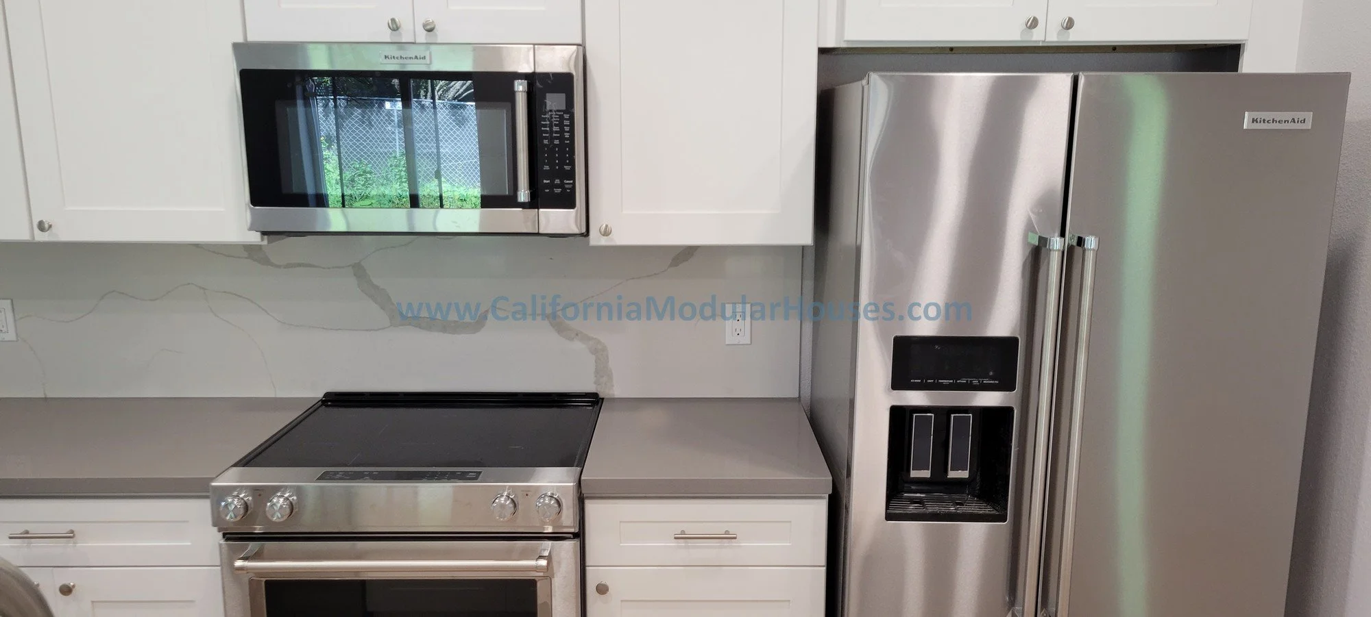 Modern kitchen with white cabinets, stainless steel microwave, stove, and refrigerator. The marble backsplash features gray veining, and a website link is visible across it.