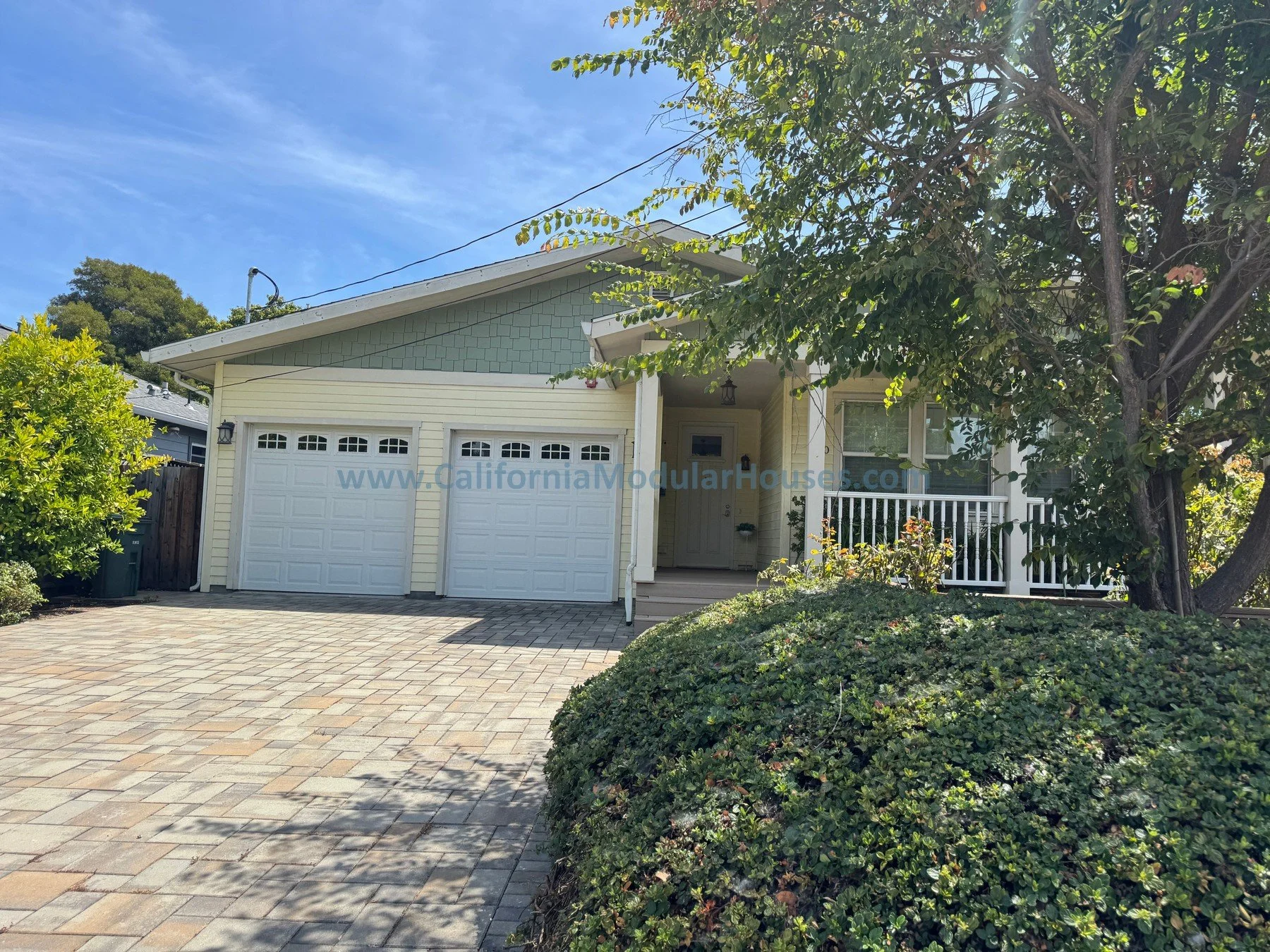 Front view of a house with a driveway made of brick pavers, a double garage door, a small porch with a white railing, and surrounding green bushes, trees, and clear blue sky.