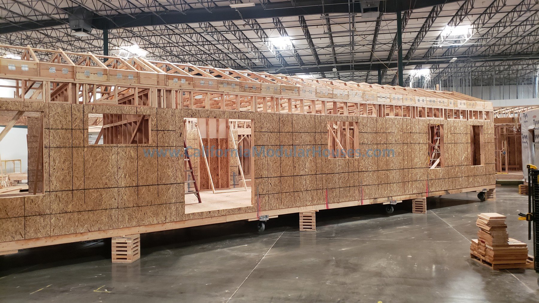 Indoor construction site of a modular tiny house with wooden framing and walls being assembled from oriented strand board panels, inside a large warehouse with exposed ceiling beams.