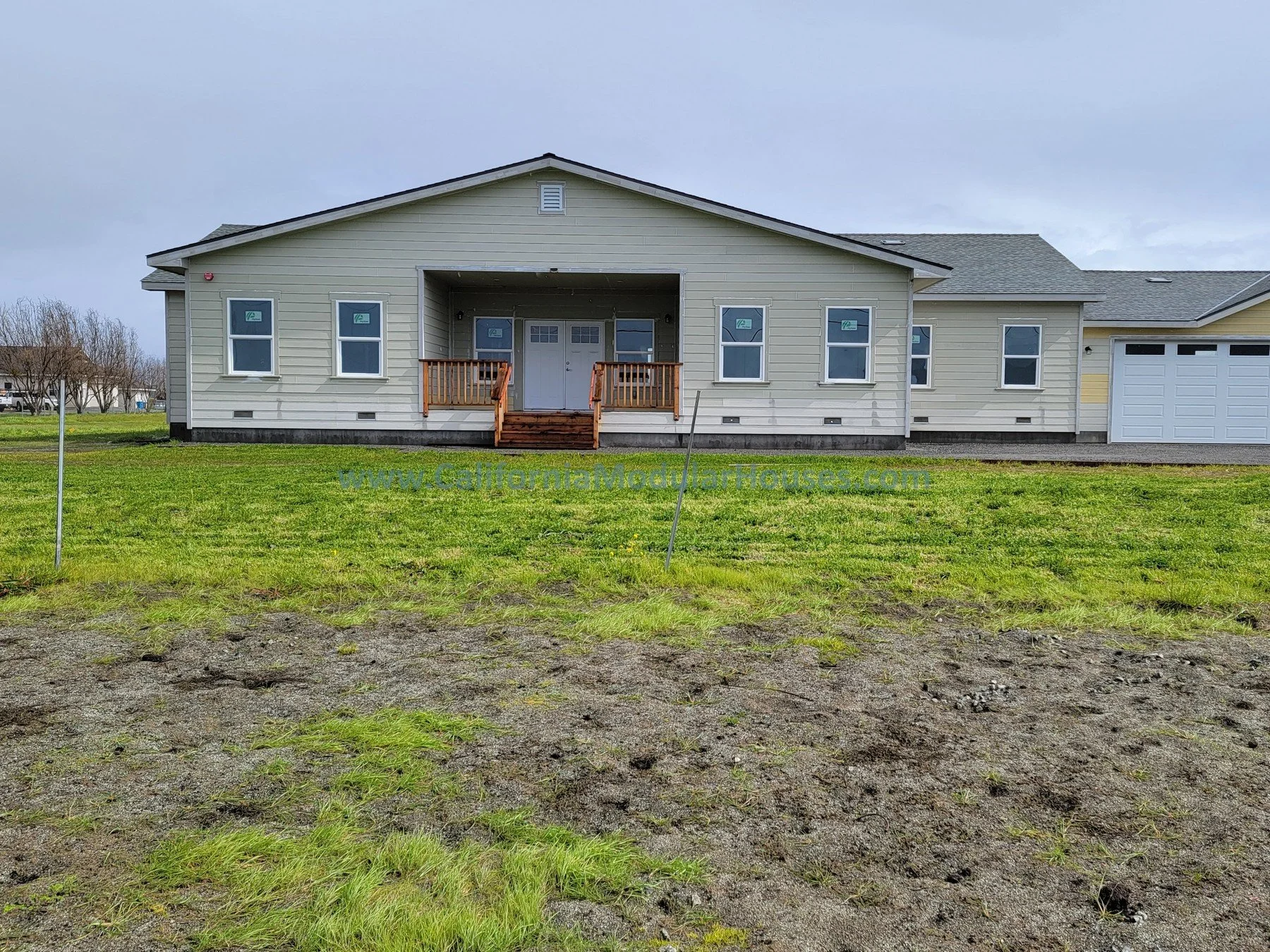 Front view of a newly built single-story house with beige siding, a large front porch with stairs, and an attached garage on the right.  Modular home.  San Juan Bautista County, CA.  Modular home.  Modular house.  Prefab.  