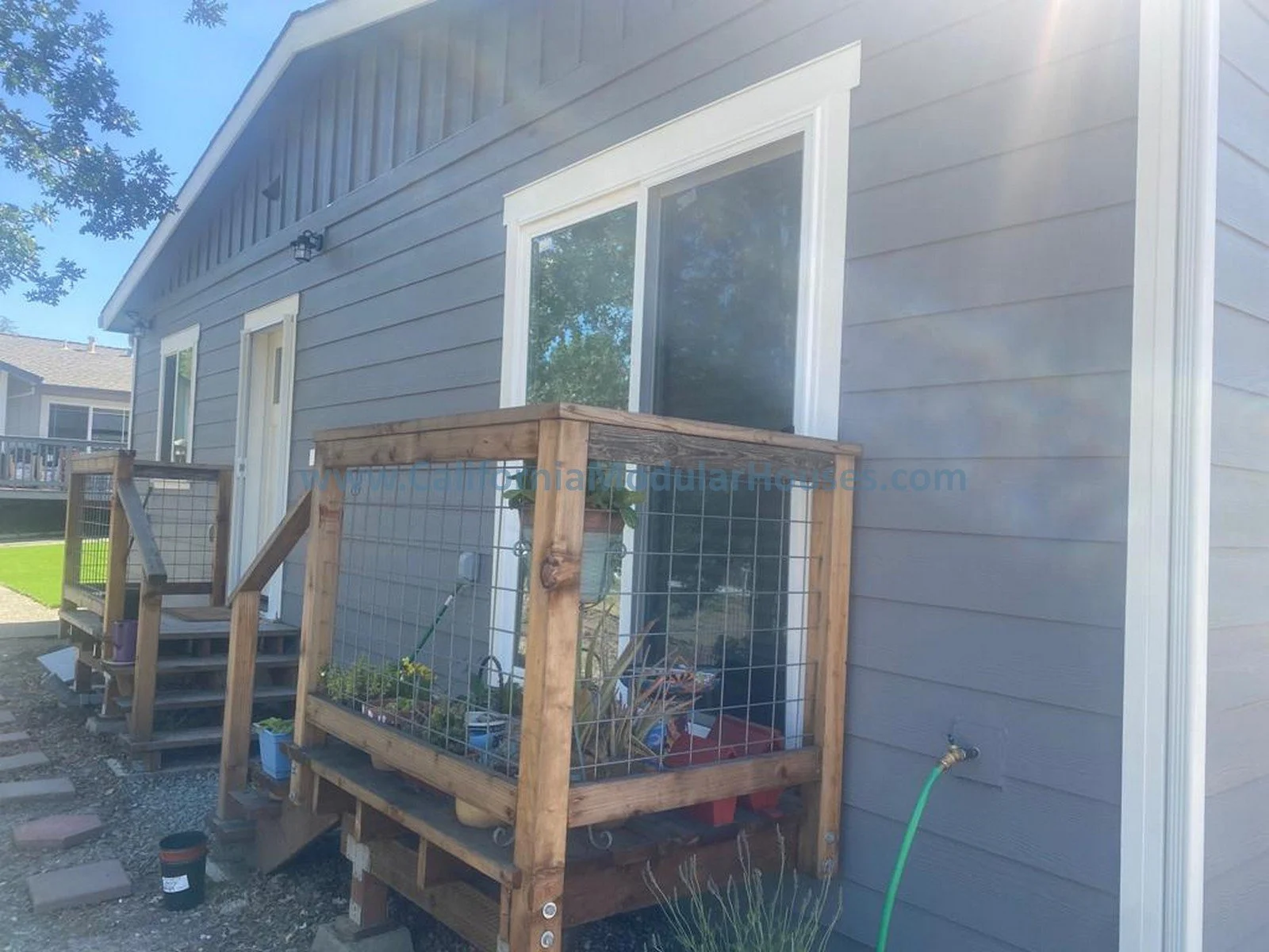 A side view of a blue house exterior with white trim, showing a small wooden raised garden bed with wire fencing and plants, a sliding glass door, and a small set of wooden stairs leading to a deck.