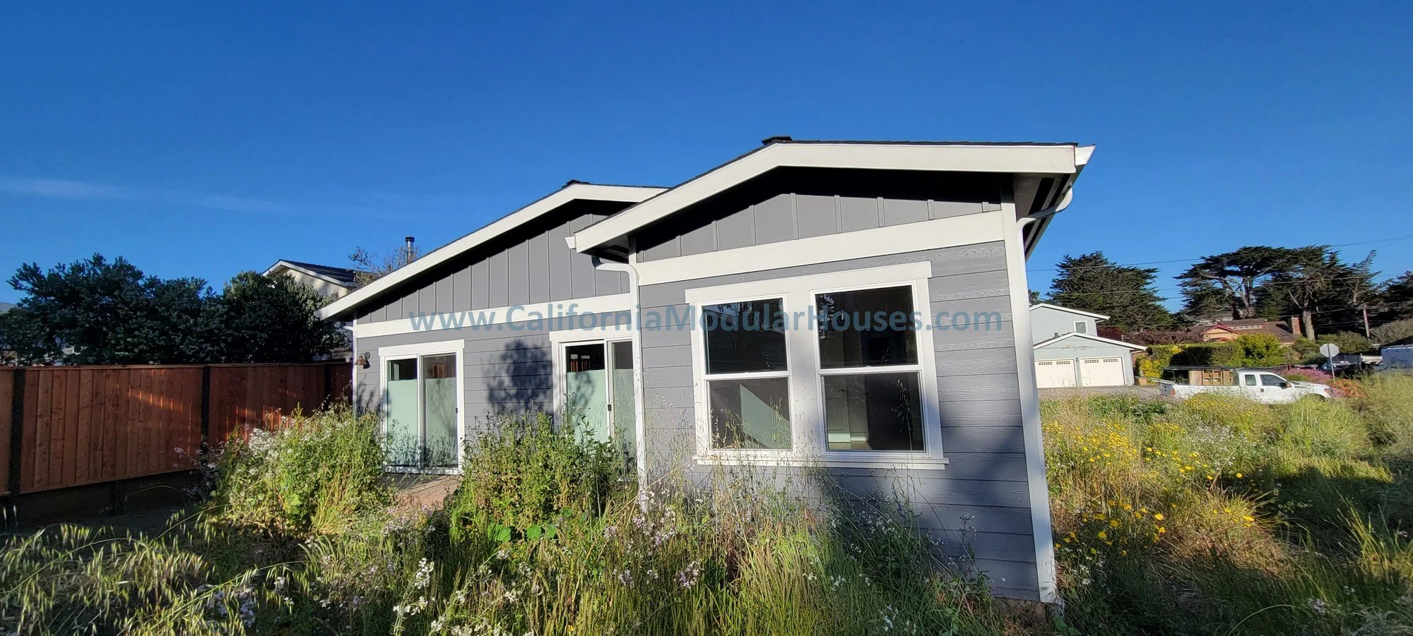 Modern gray house with white trim, large windows, and a landscaped front yard with wildflowers and tall grass. Clear blue sky in the background.