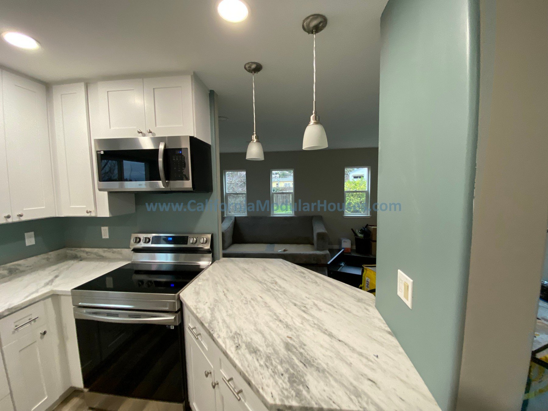 Prefab modular ADU Modular Granny Flat A modern kitchen with white cabinets, stainless steel microwave and oven, and marble countertops. Two pendant lights hang from the ceiling, and the kitchen opens into a living area.