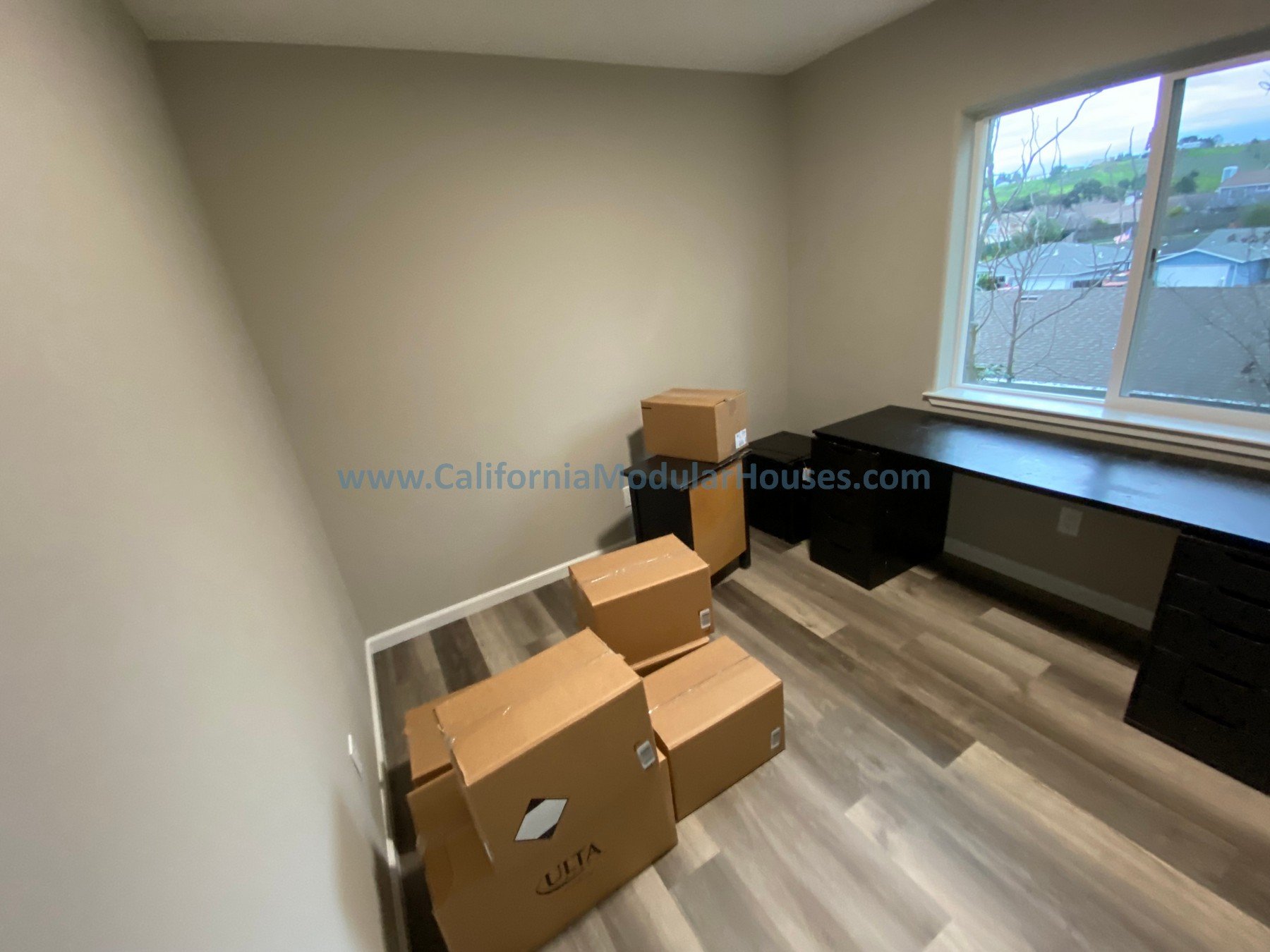 Empty room with four cardboard boxes on the floor, a black desk under a window, and a view of a suburban neighborhood through the window.  Modular Accessory Dwelling Unit Modular ADU