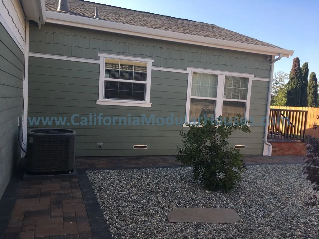 Backyard of a house with green siding, white trim, and two windows. There's a small tree in a gravel area, a brick patio, and an air conditioning unit on the left side.