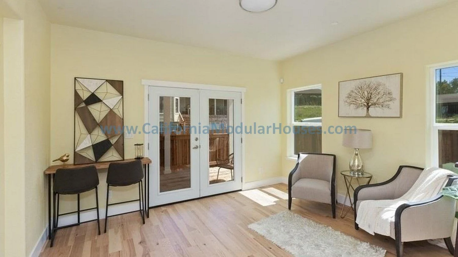 PreFab Modular for California.  Living room with a pair of armchairs, a side table with a lamp, and multiple windows; French doors leading outside; artwork on the walls; hardwood floors.  Modular house interior.  
