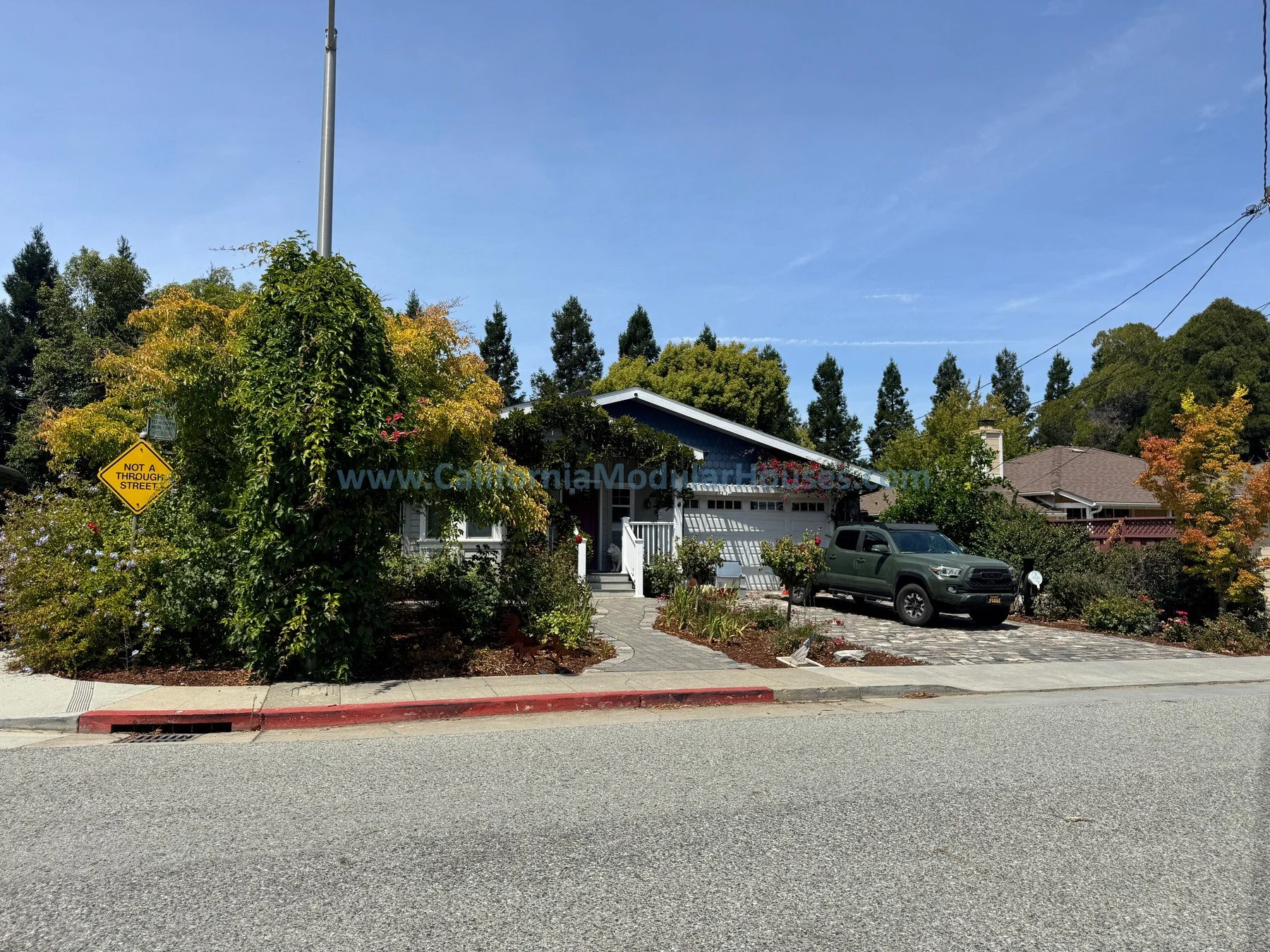 A house with a front porch and garage, surrounded by landscaped garden with bushes and trees, parked gray truck, parked car, and a street with a red curb. There is a yellow sign that reads 'Not a Through Street' and a sidewalk.