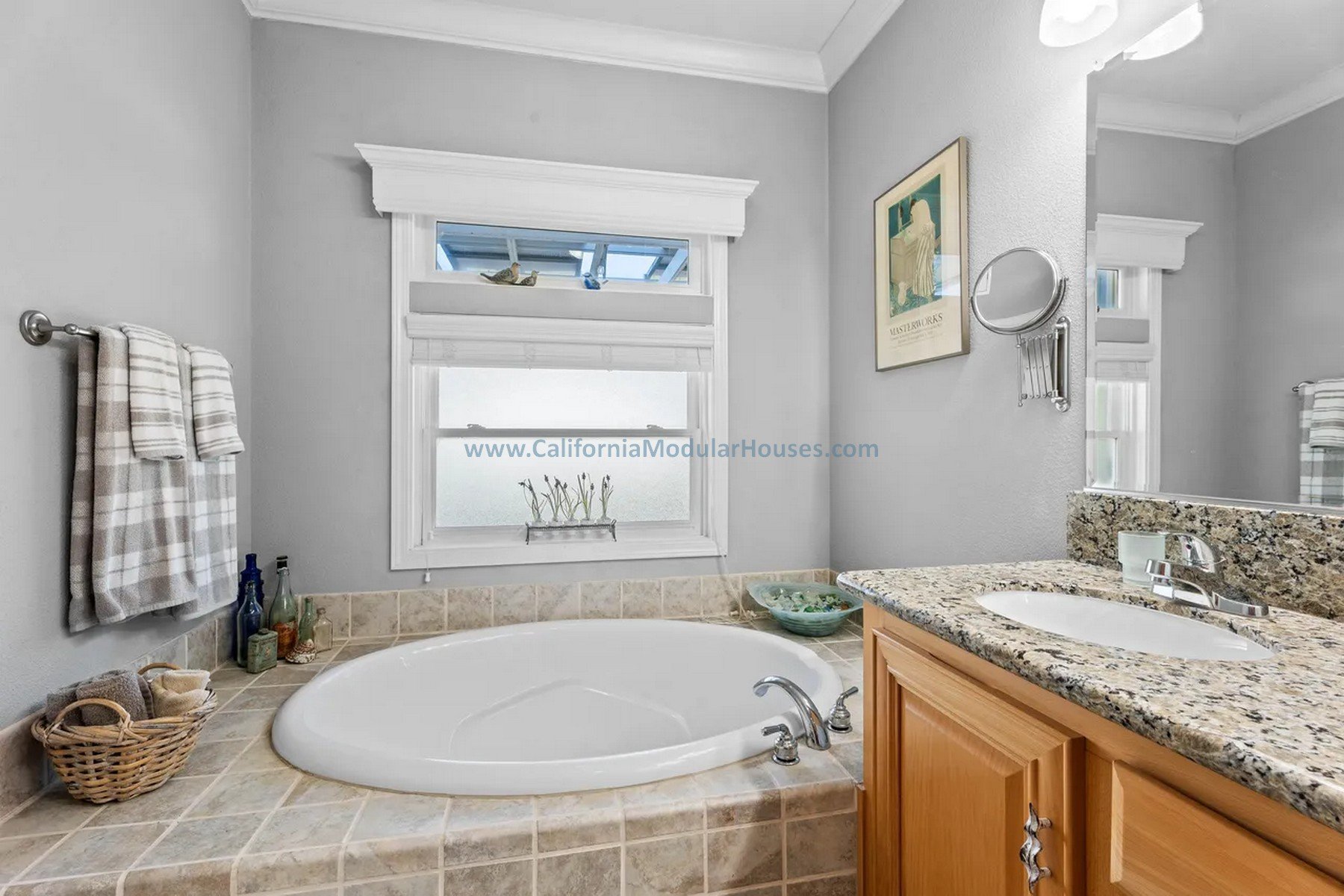 A bathroom featuring a built-in bathtub with beige tiled surround, a window with white trim, a granite countertop with sink, a mirror, and a wall-mounted magnifying mirror. Prefab Modular Single Family Residence. California. Cape Cod home with attic