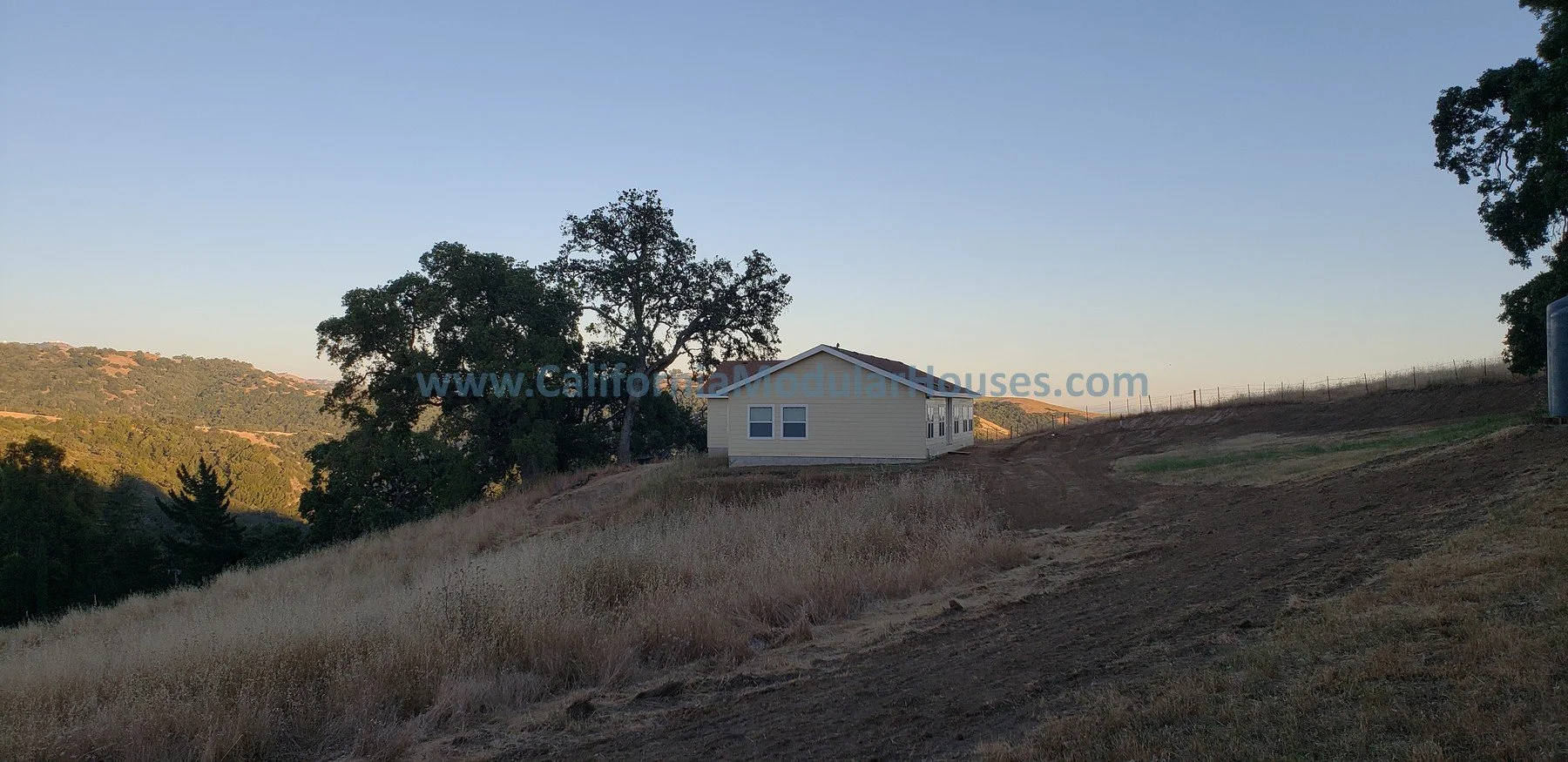 A small house on a hilltop with grass and dirt roads, a large tree to the left, and distant hills in the background, with a blue sky.  California Modular Houses, Bay Area Prefab Modular Home, Modular Home CA,