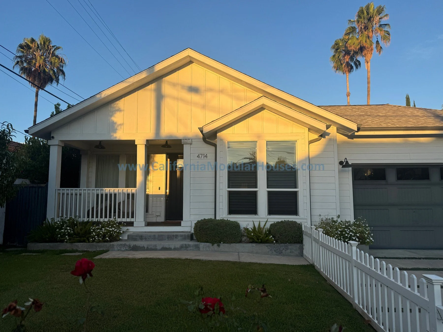 Front view of a white house with a small porch and a garden, palm trees in the background, and a blue sky at sunset.