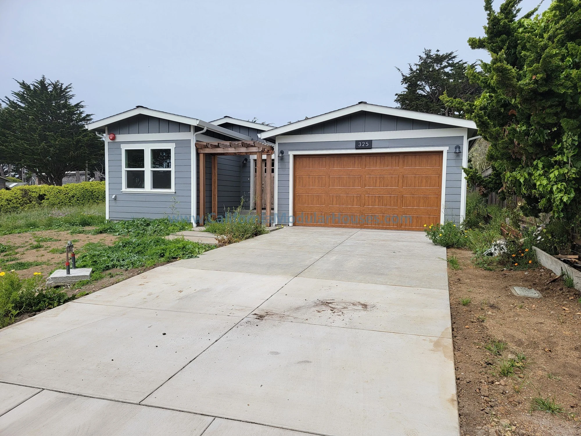 Front view of a modern house with a double garage door, painted in blue, and a small front yard with some greenery. The driveway is concrete, leading to the garage, and there is a wooden pergola structure near the entrance.