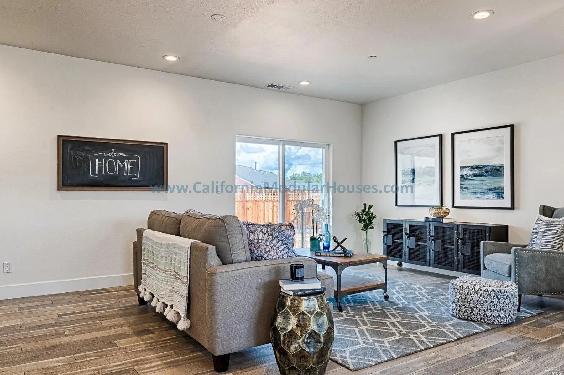 Living room with a beige sofa, gray armchair, black sideboard, wooden coffee table, patterned pouf, decorative items, framed artwork, and a window showing a backyard with a wooden fence and cloudy sky.