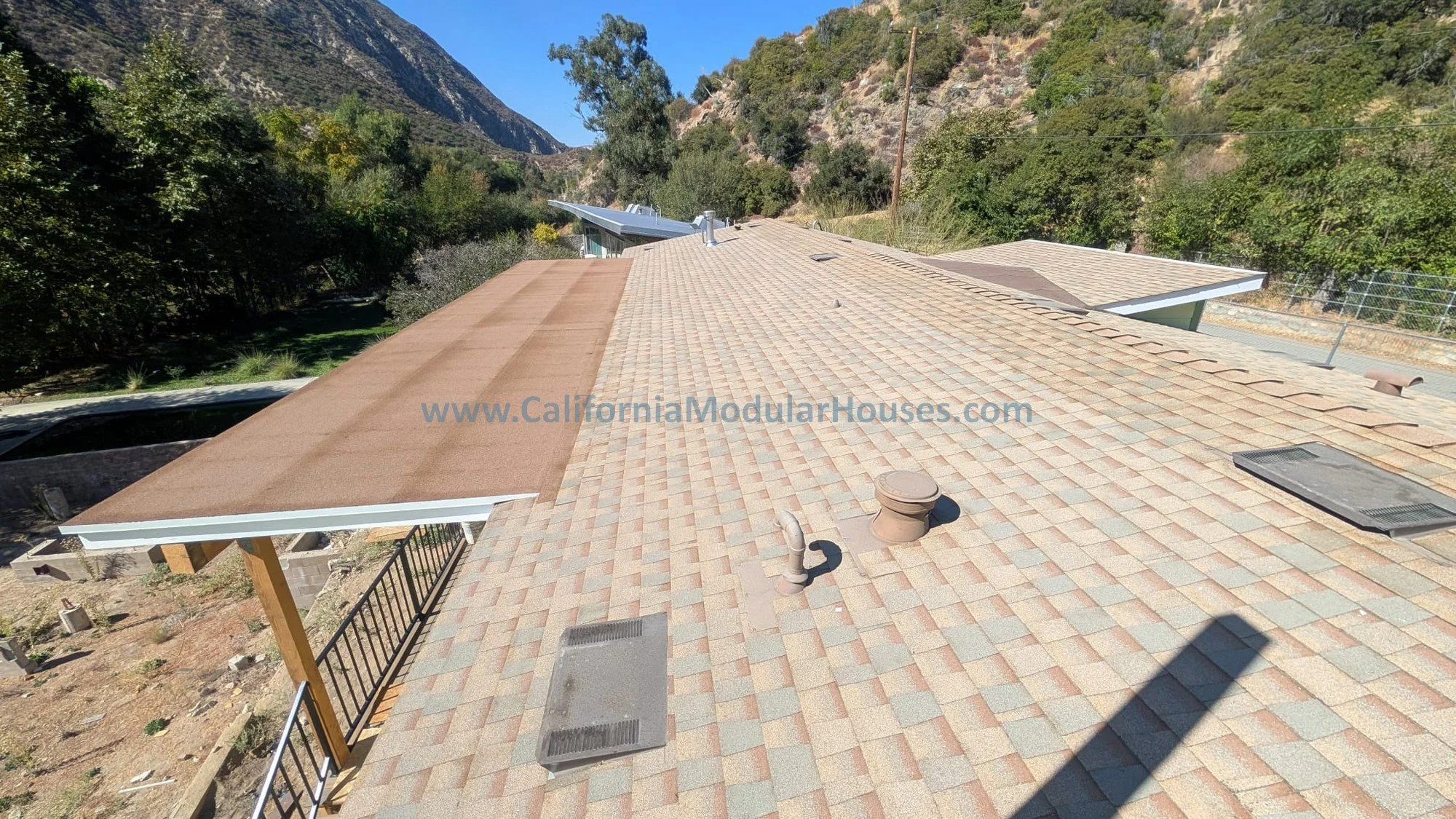A rooftop with a mix of new and old roof sections, the newer section covered with brown asphalt shingles and the older section with weathered brown shingles. The rooftop has vents, a skylight, and a small ladder near the edge. Surrounding landscape i