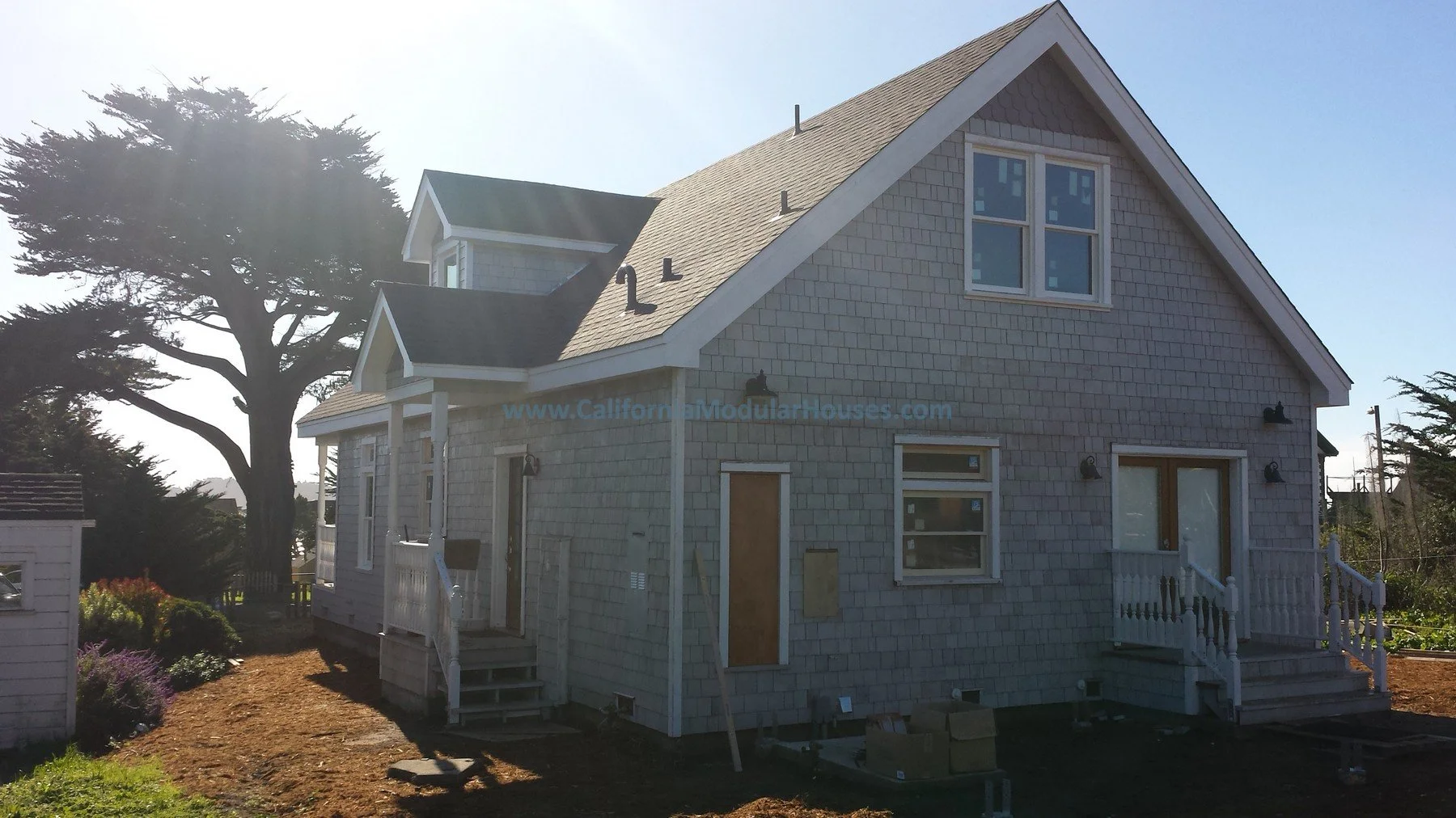 A two-story house under construction with grey shingles exterior, multiple windows, stairs leading to doors, and a large tree in the background.  Custom design coastal commission cape cod modular home.  Pre-fab modulars.  California.