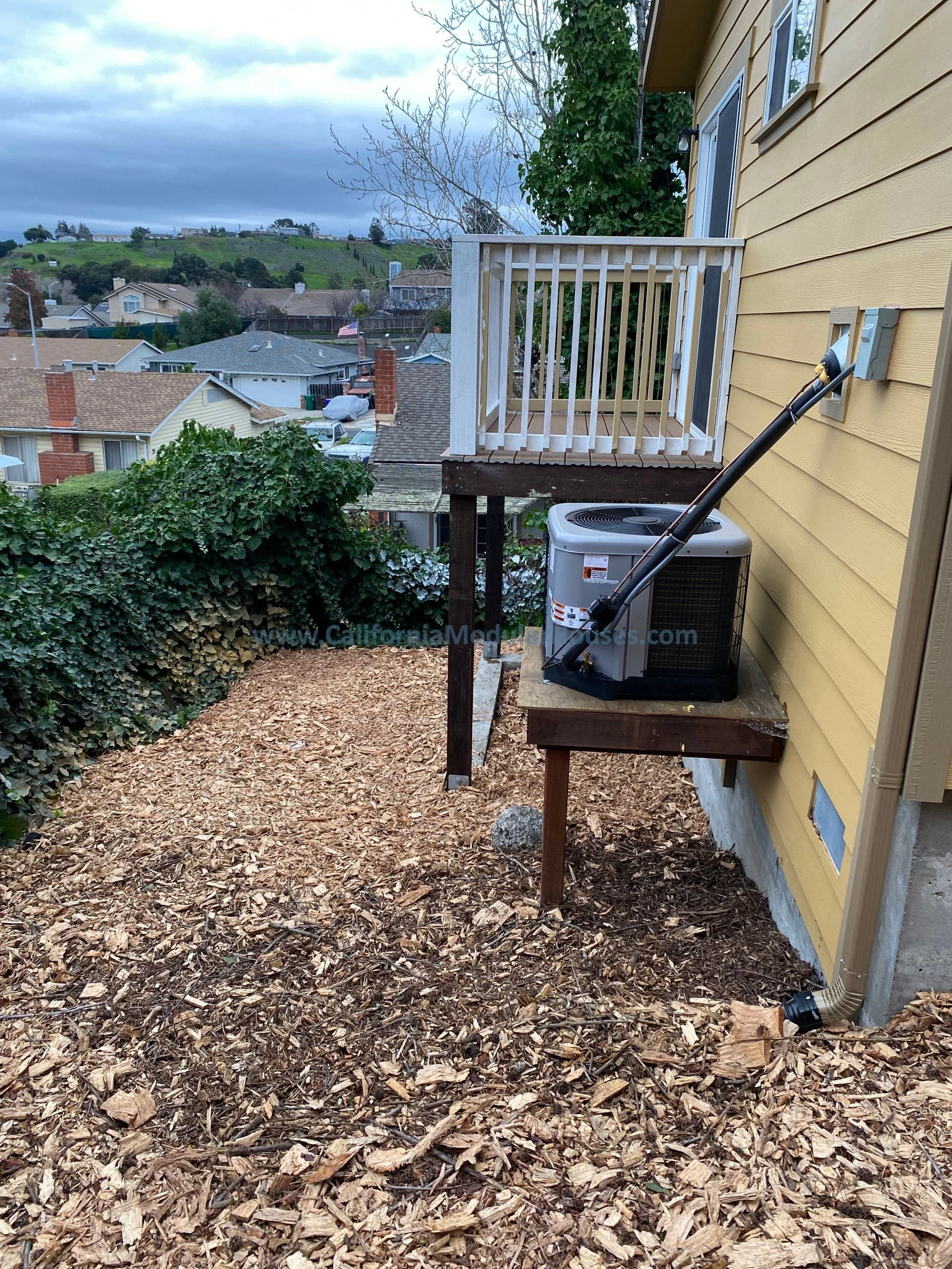 Backyard with a small wooden deck, an air conditioning unit, and a yellow house exterior. The ground is covered with wood mulch, and there is a view of a neighborhood with houses and hills in the distance.  Modular Granny Flat  Modular Accessory Dwel