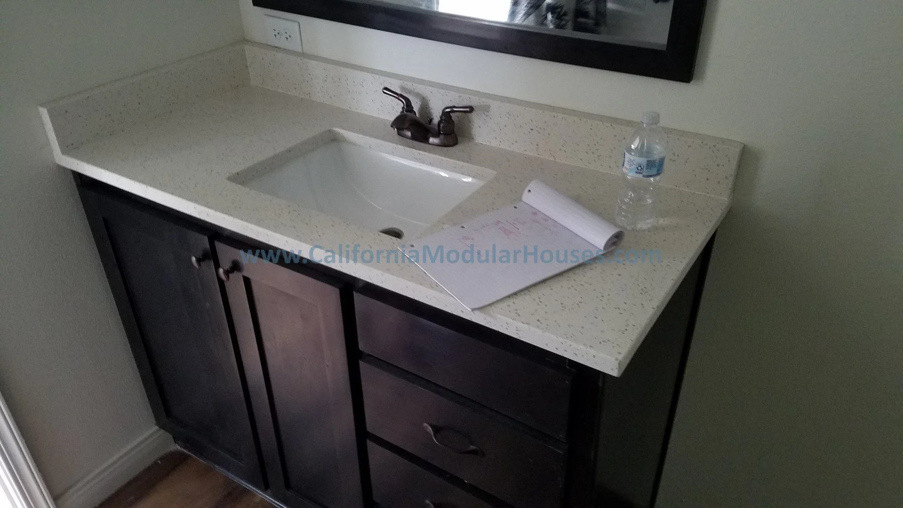 A bathroom sink with a black cabinet and a speckled white countertop, a faucet, a bottle of water, and a notepad.  California Modular Home,  California Modular, Prefab Modular Homes California, Modular Home CA,